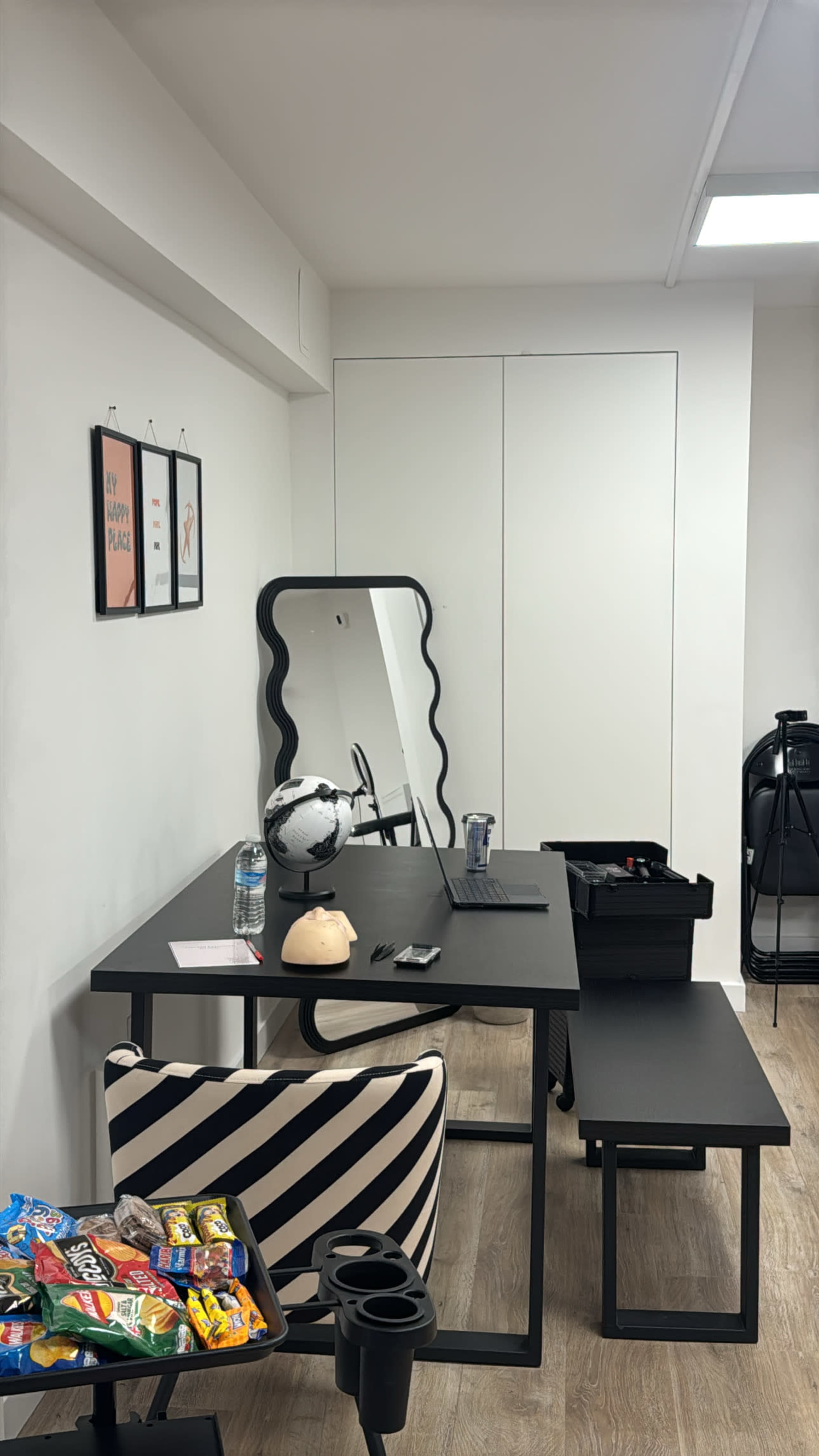 The image shows a modern workspace with a black desk, a large mirror, a chair with a striped pattern, and a small table, all positioned against a white wall with some framed artwork.
