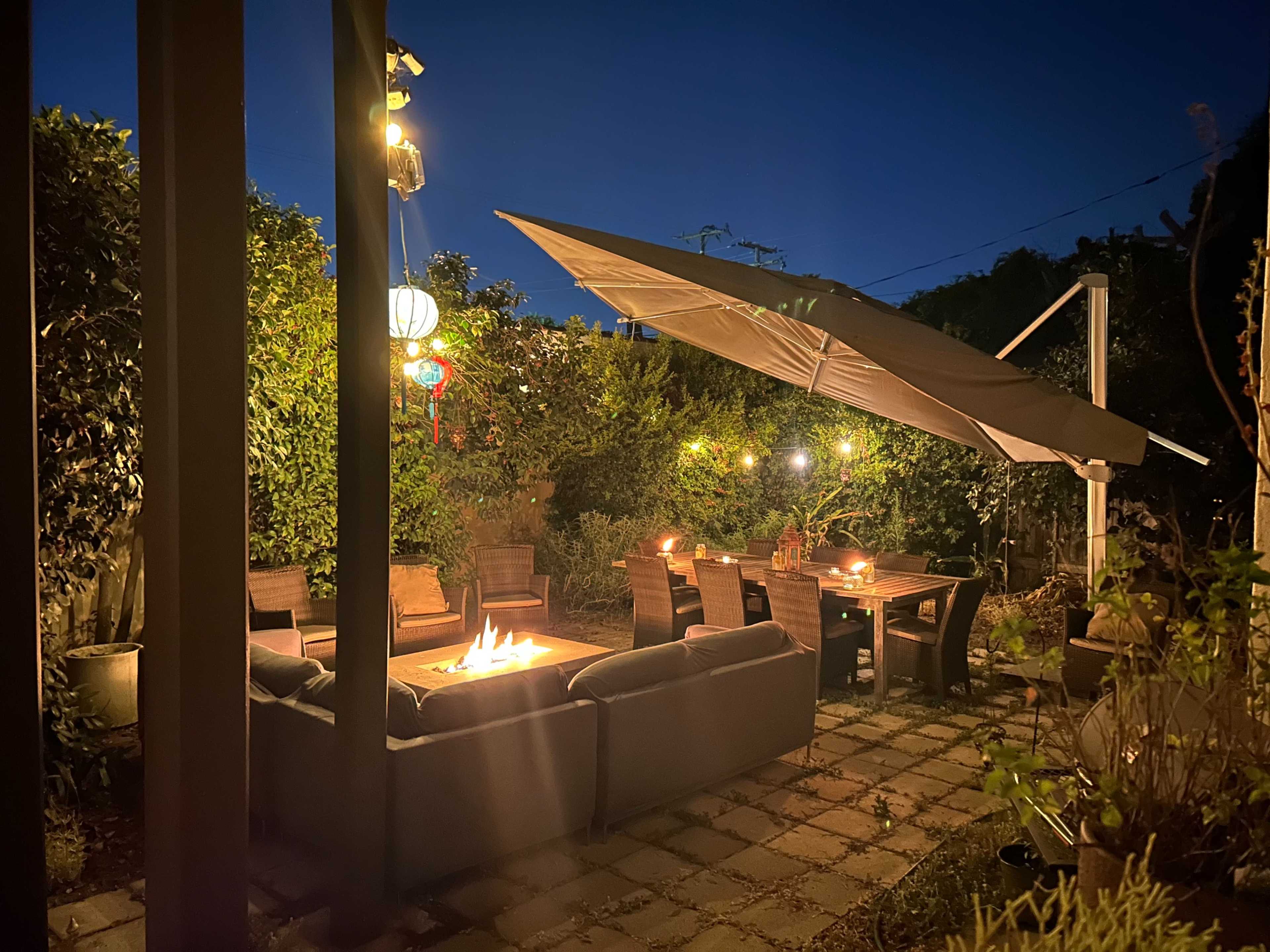 A patio area features a seating arrangement with a fire pit and an umbrella, illuminated by ambient lights and surrounded by greenery at night.