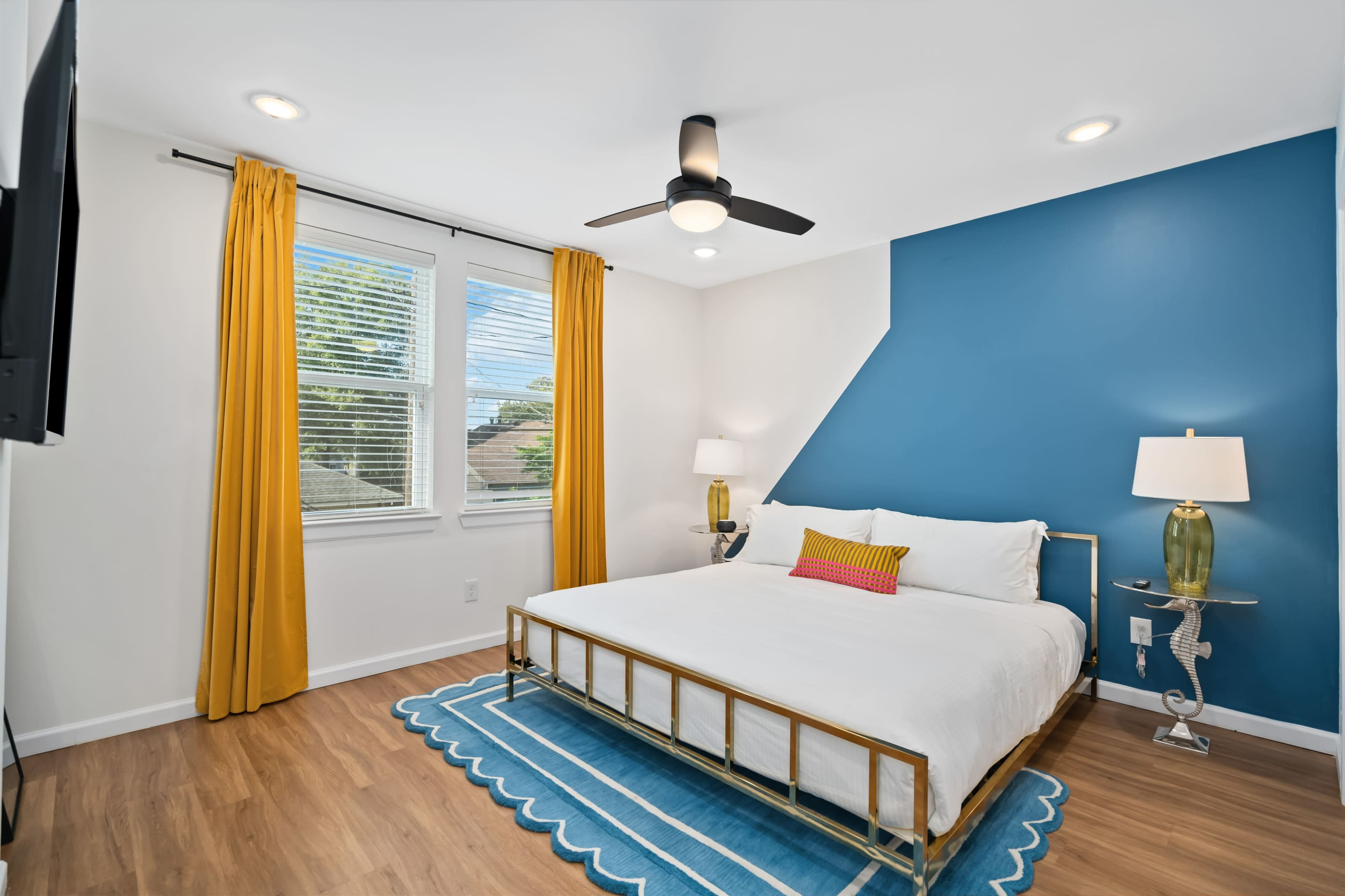 A modern bedroom with a white bed, yellow curtains, a blue accent wall, and hardwood flooring.