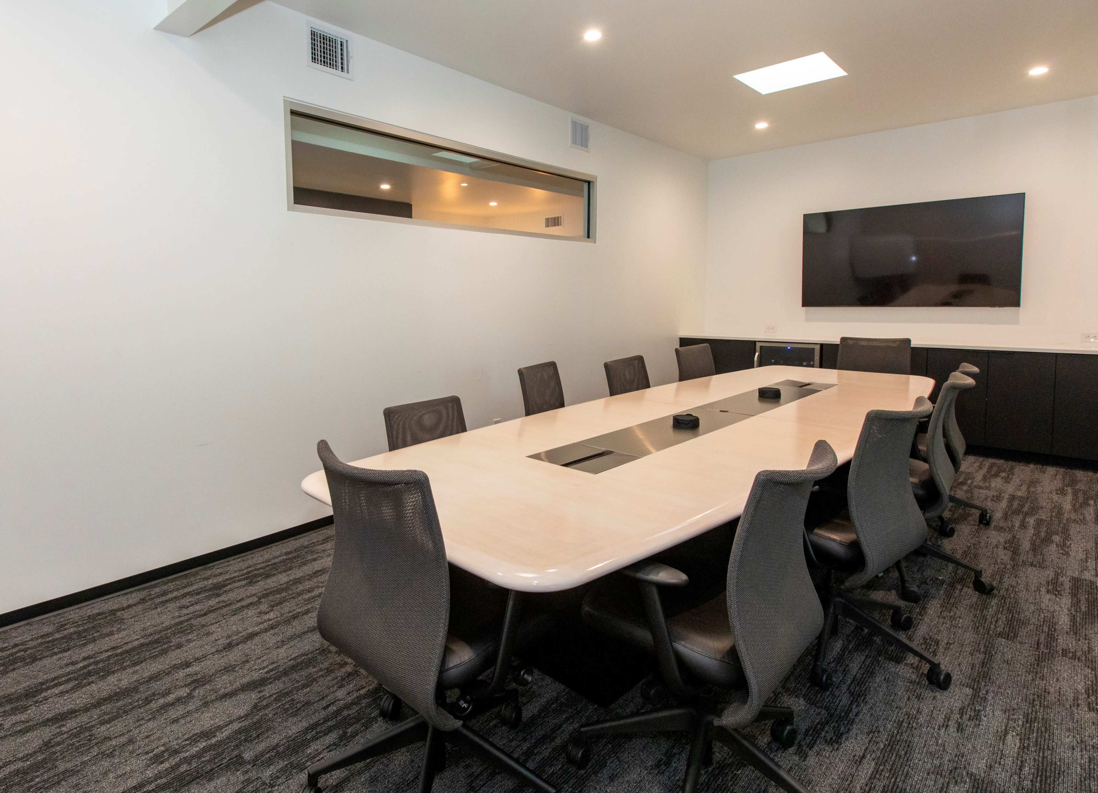 A modern conference room features a long oval table surrounded by multiple black ergonomic chairs, with a wall-mounted TV and a window providing natural light.