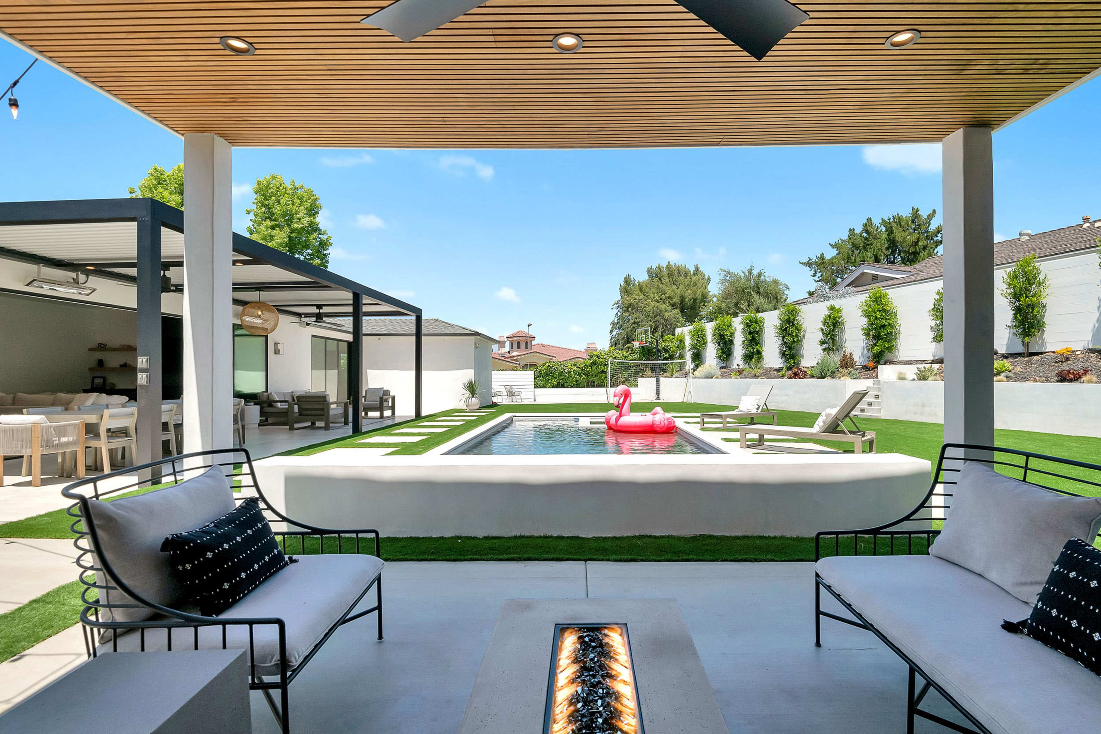 A modern outdoor living space features a rectangular pool with a pink inflatable flamingo, surrounded by seating areas and a landscaped yard.