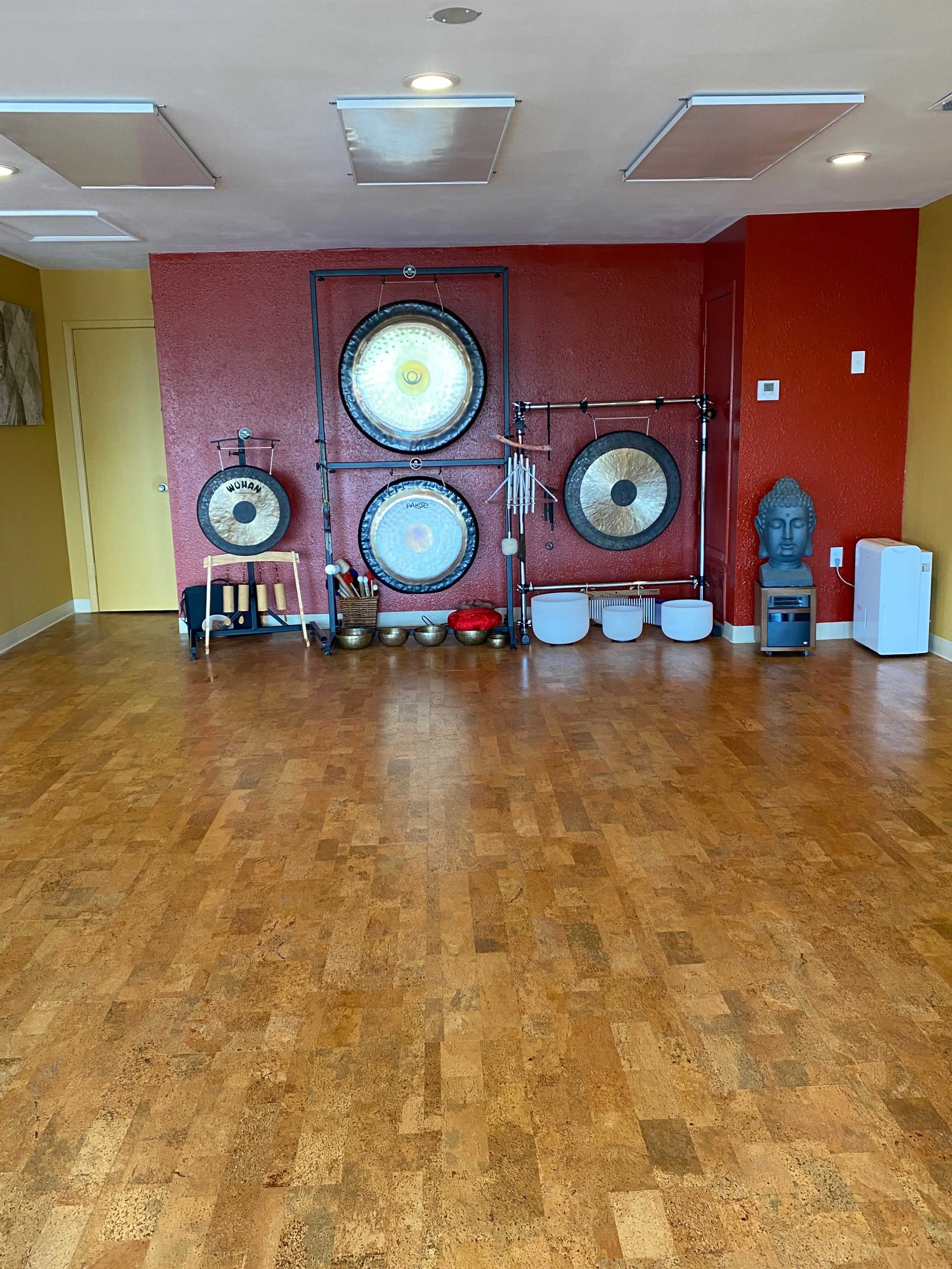 The image shows a meditation room featuring large gongs mounted on the wall, various musical instruments, and a statue of a Buddha, with a polished wooden floor and walls painted in warm colors.