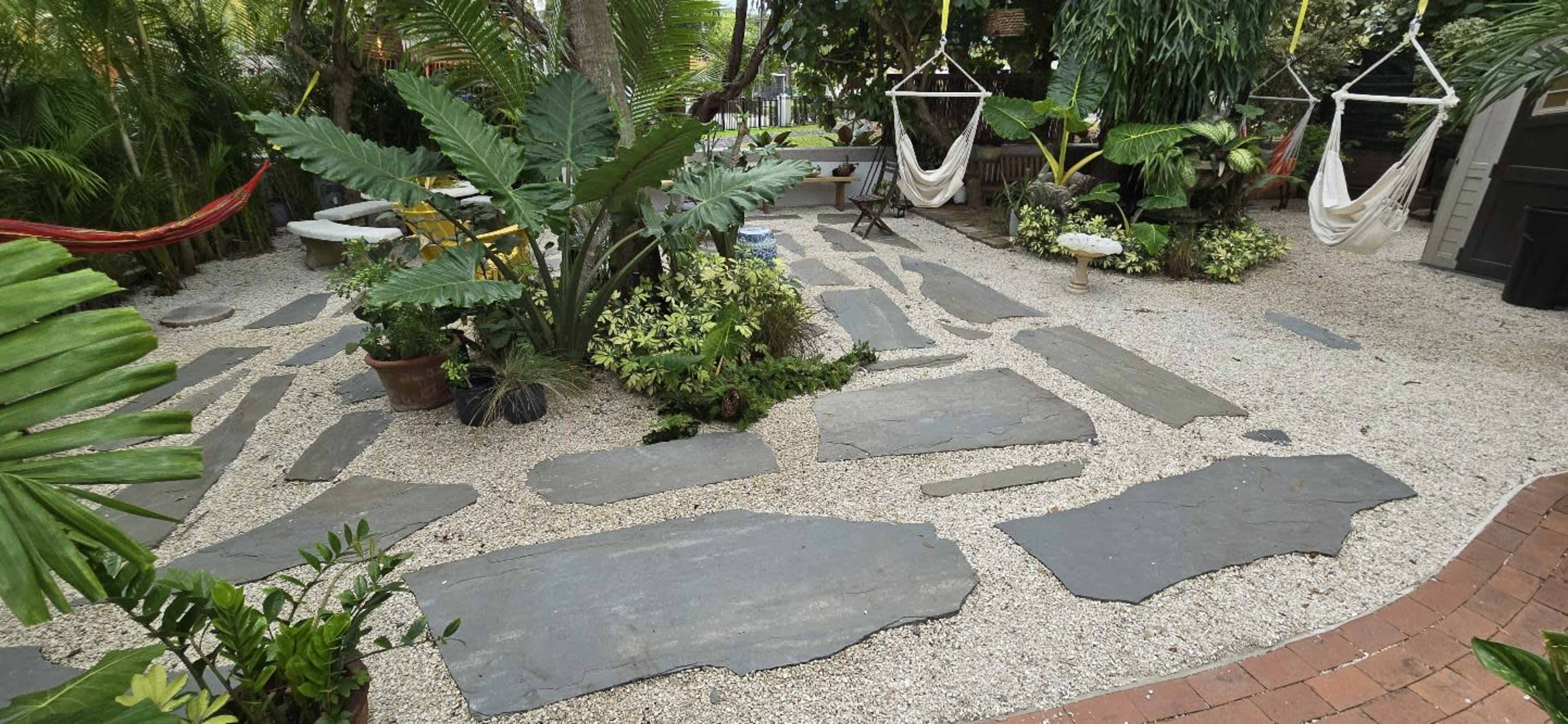 The image shows a landscaped outdoor area featuring a stone pathway surrounded by various plants, hammocks, and decorative elements.