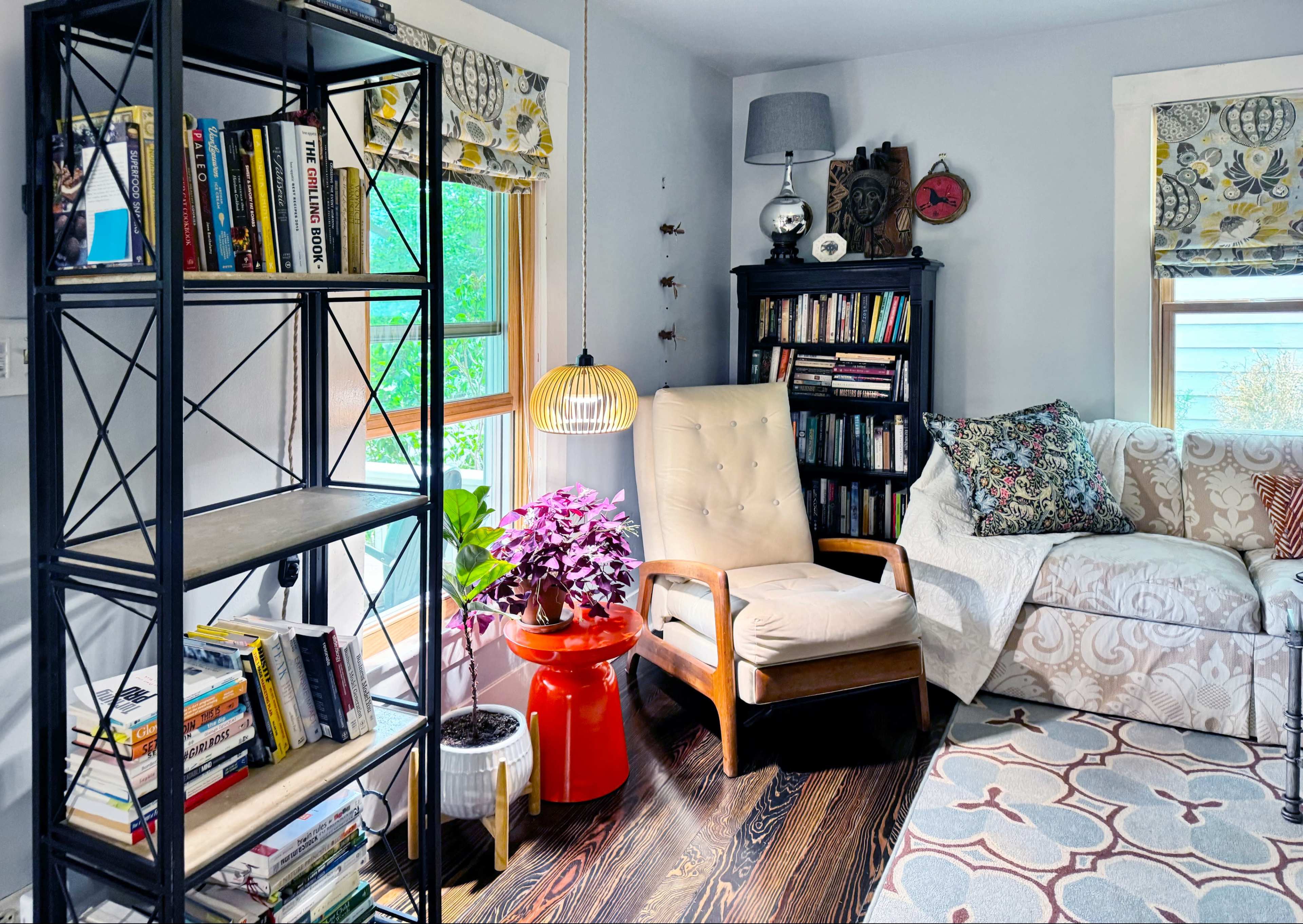 The image shows a cozy corner of a room featuring a bookshelf, a comfortable chair, a small side table, and a sofa, all illuminated by natural light coming through a window.