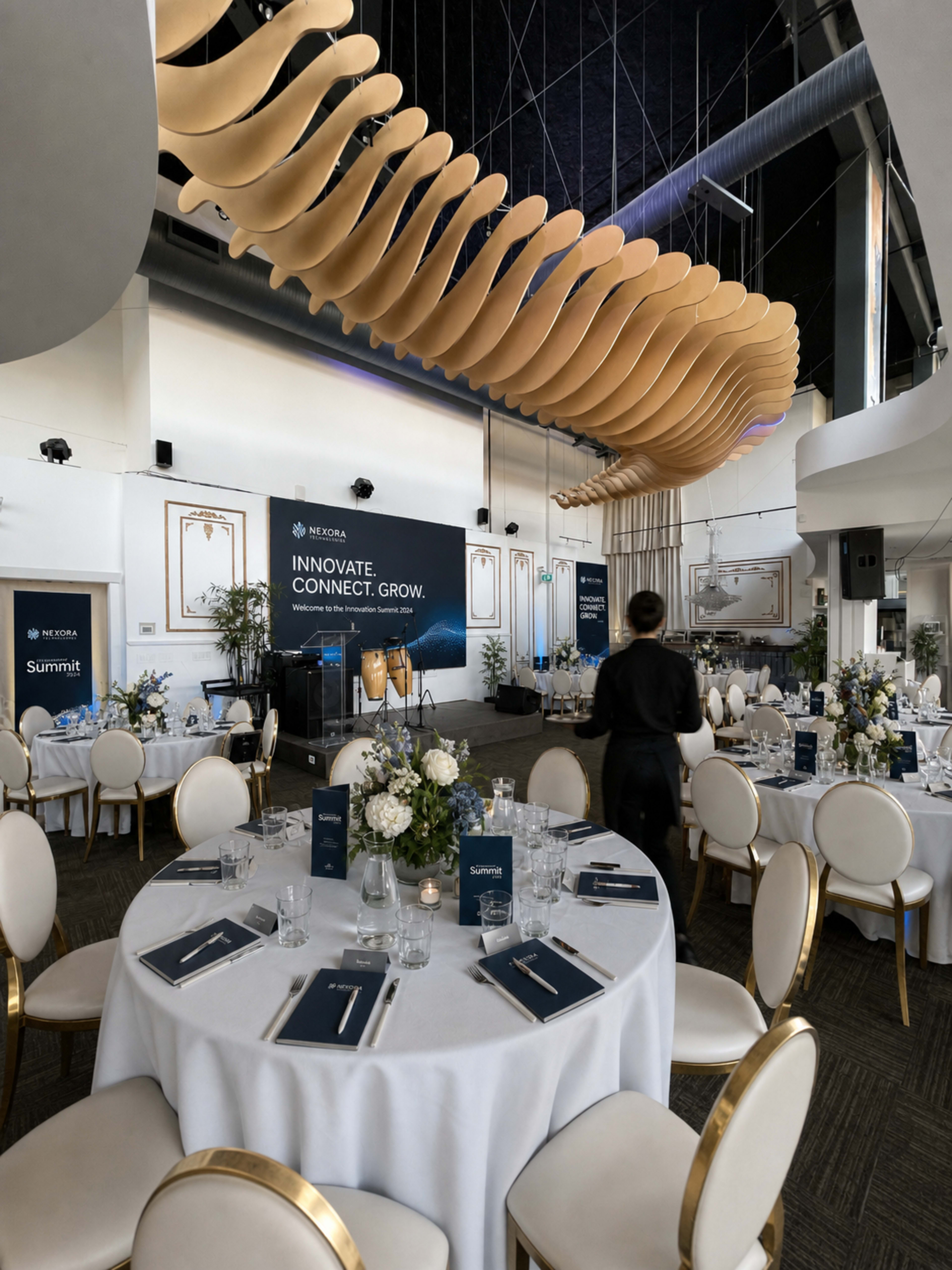 A spacious banquet room features elegantly set round tables with white linen, centerpieces of flowers, and a backdrop displaying the words "INNOVATE. CONNECT. GROW."