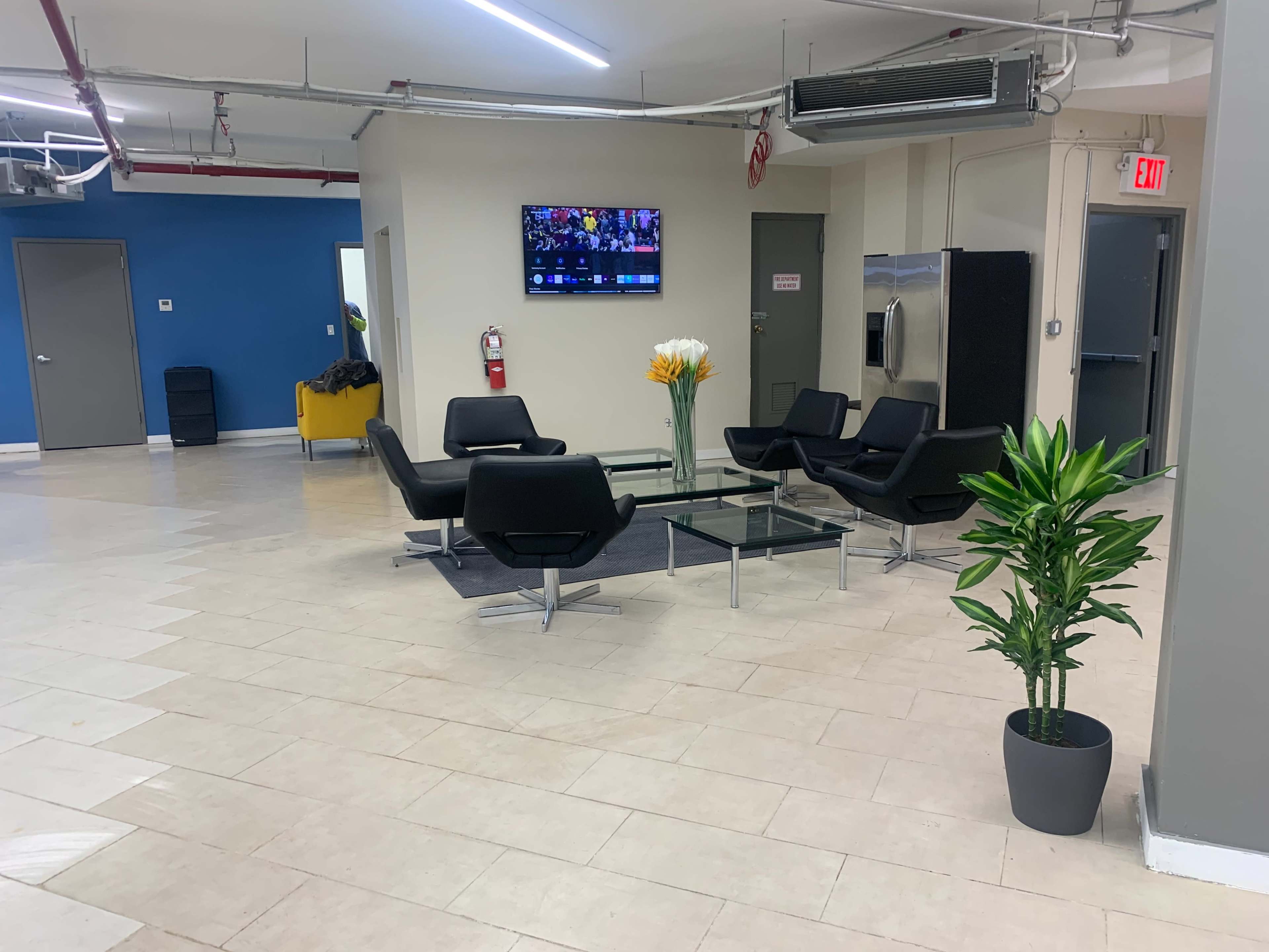 The scene features a modern waiting area with black chairs surrounding a glass table, a potted plant, and a television mounted on the wall.