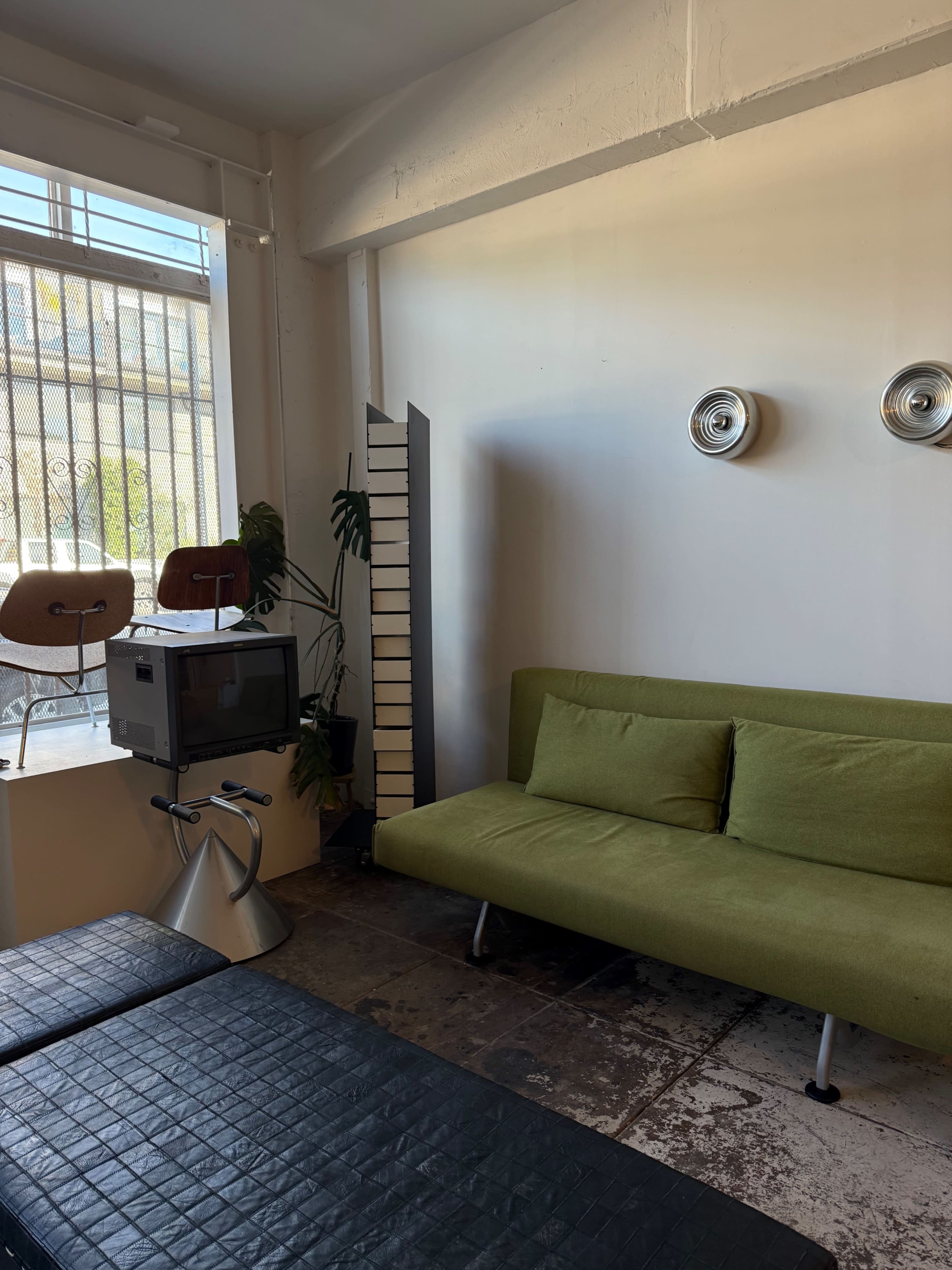 The image shows a minimalist living space featuring a green sofa, a vintage television, and a backdrop of a large window with plants and wooden chairs in the corner.