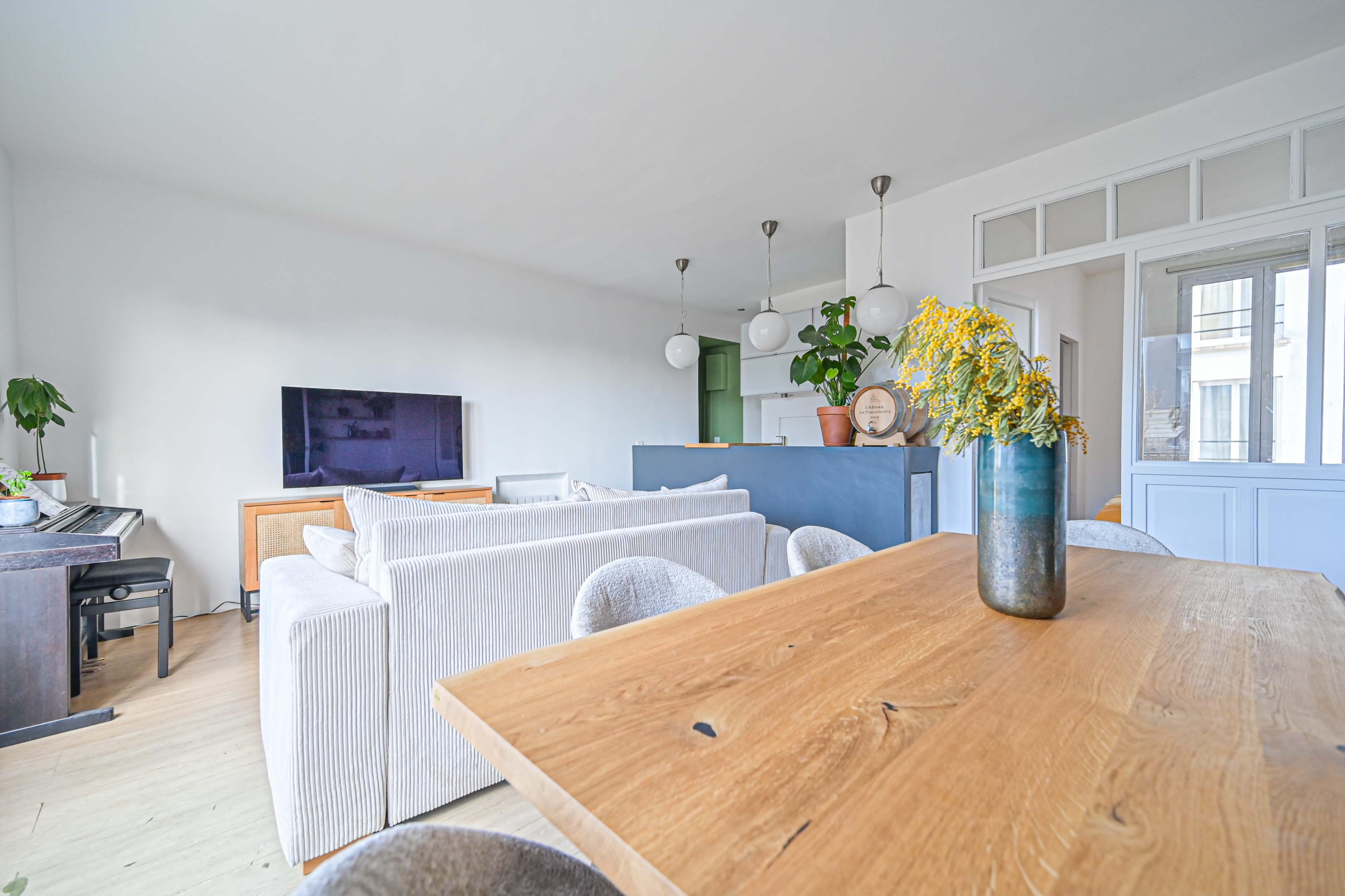 The image shows a modern living space featuring a light-colored sofa, a wooden dining table with flower arrangements, and a view of a kitchen area in the background.
