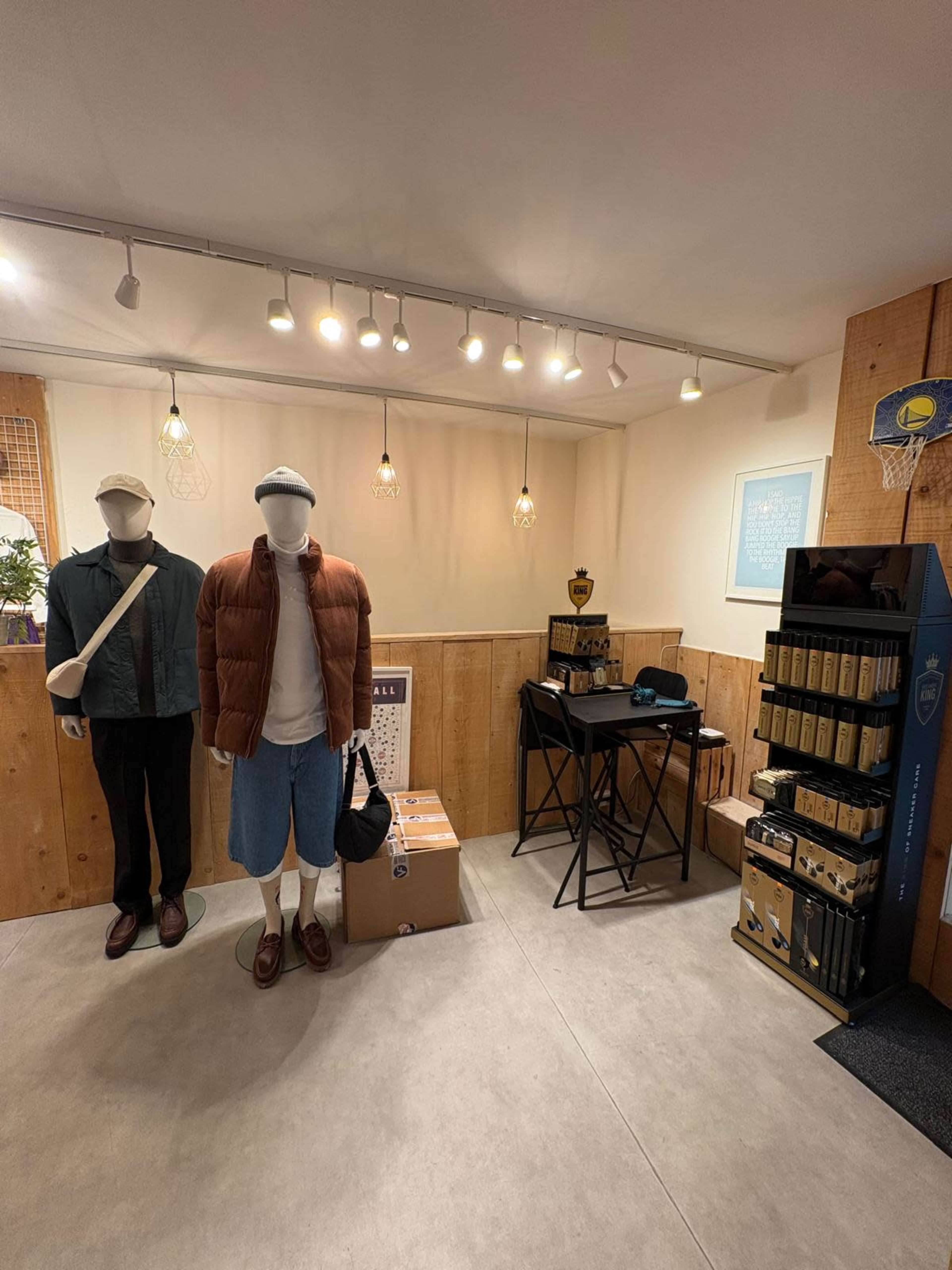 The image shows a retail space featuring two mannequins dressed in casual clothing and a display table with products nearby, all against a wooden backdrop.