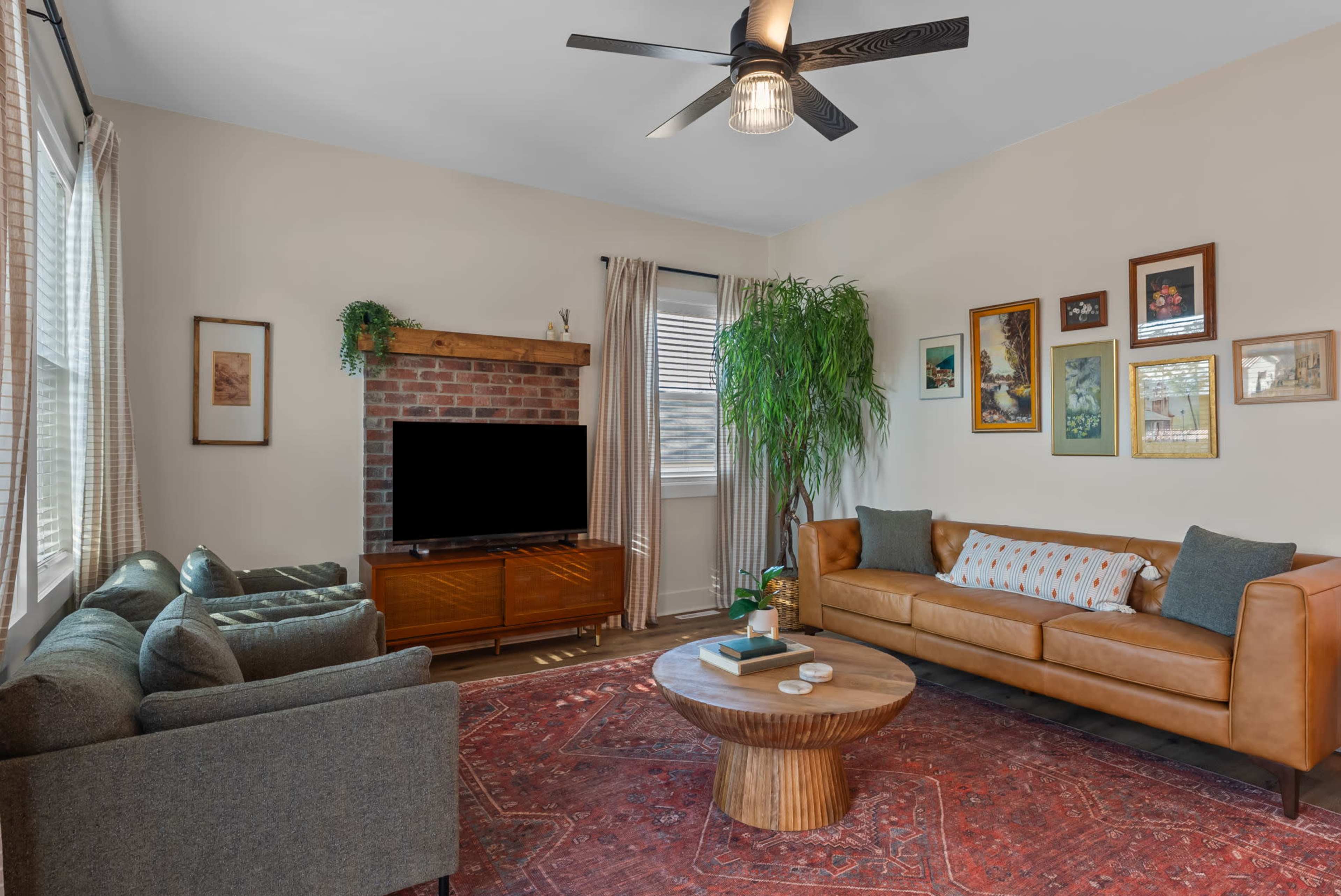 The living room features a brick fireplace, a brown leather sofa, two gray armchairs, a circular wooden coffee table, and a large television on a wooden console.