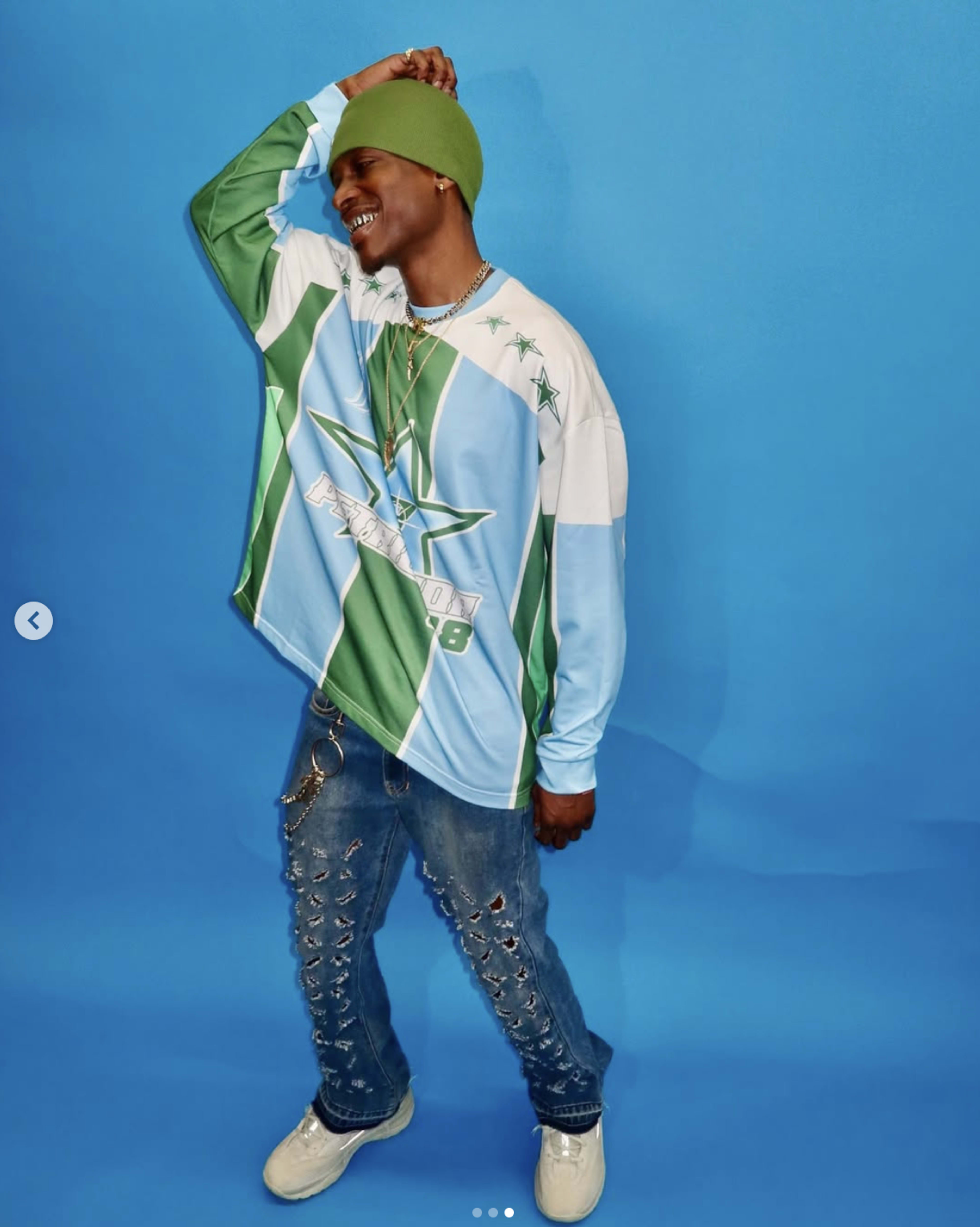 A person wearing a colorful oversized shirt and green beanie poses against a blue background.