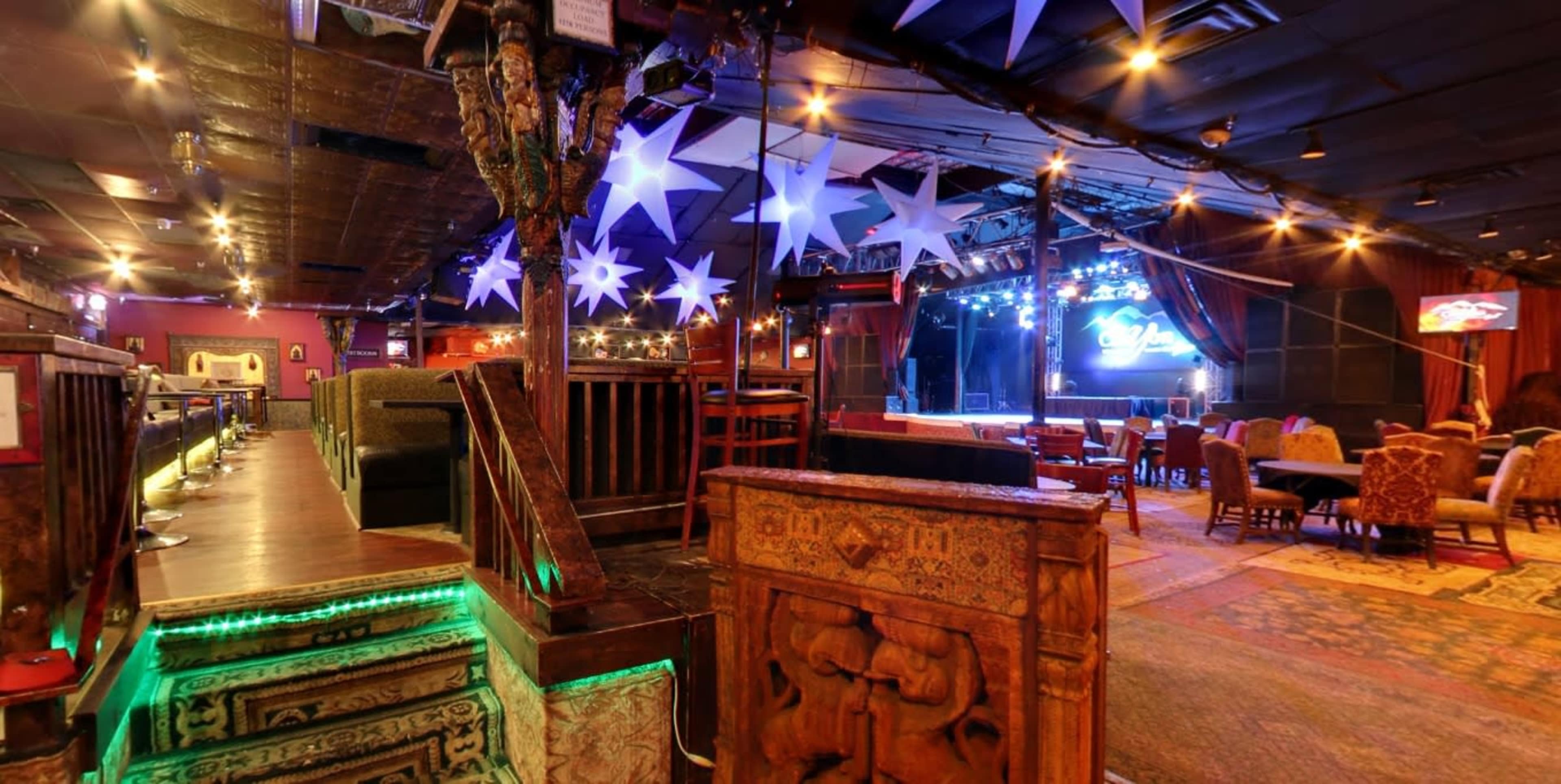 The interior of a vibrant live music venue features a stage with blue lights, colorful decor, and a mix of seating arrangements.