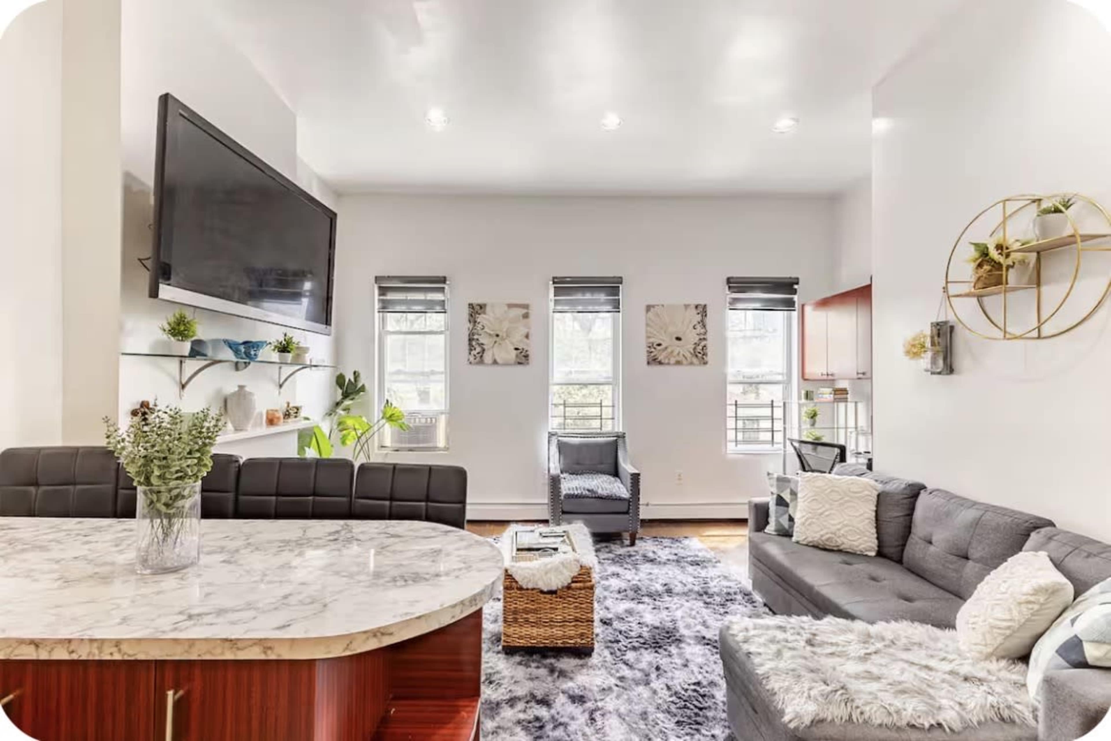 The image shows a modern living room with a sectional sofa, a marble countertop, a television mounted on the wall, and decorative plants.