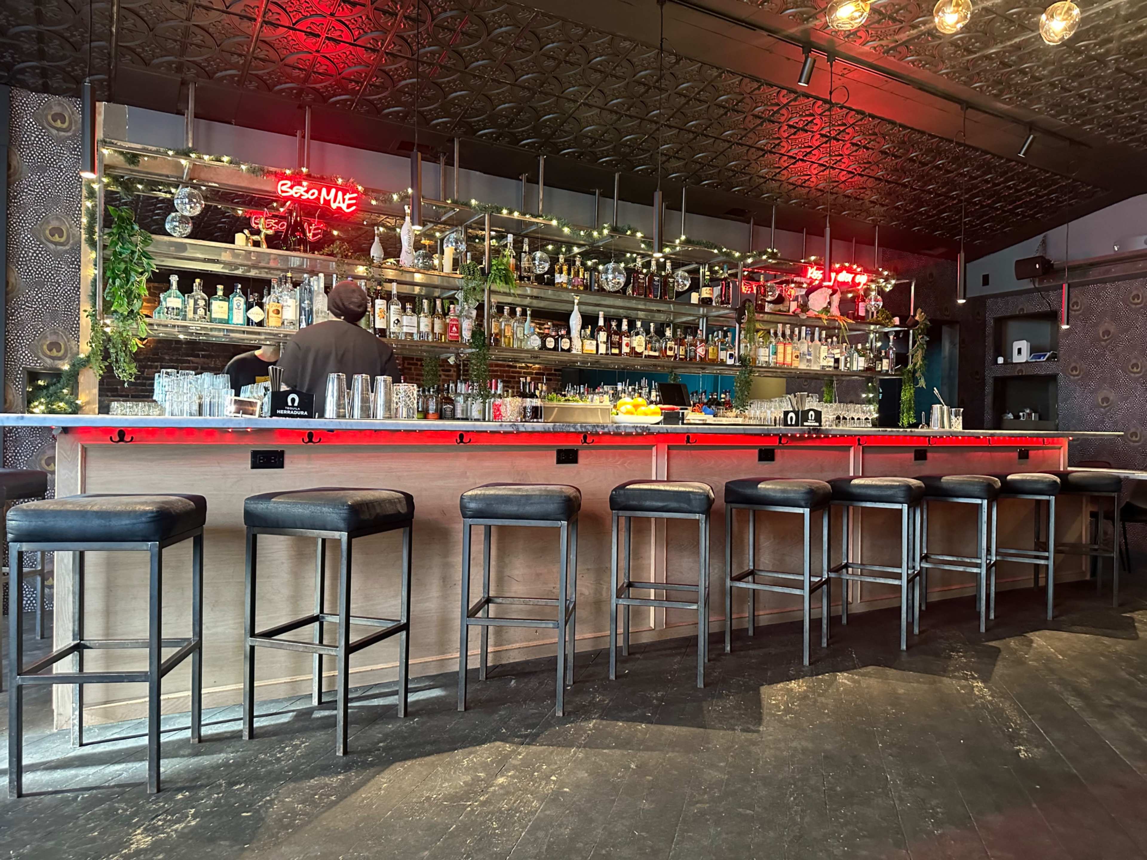 A dimly lit bar features a long counter with several bar stools, shelves stocked with various bottles, and a bartender preparing drinks.