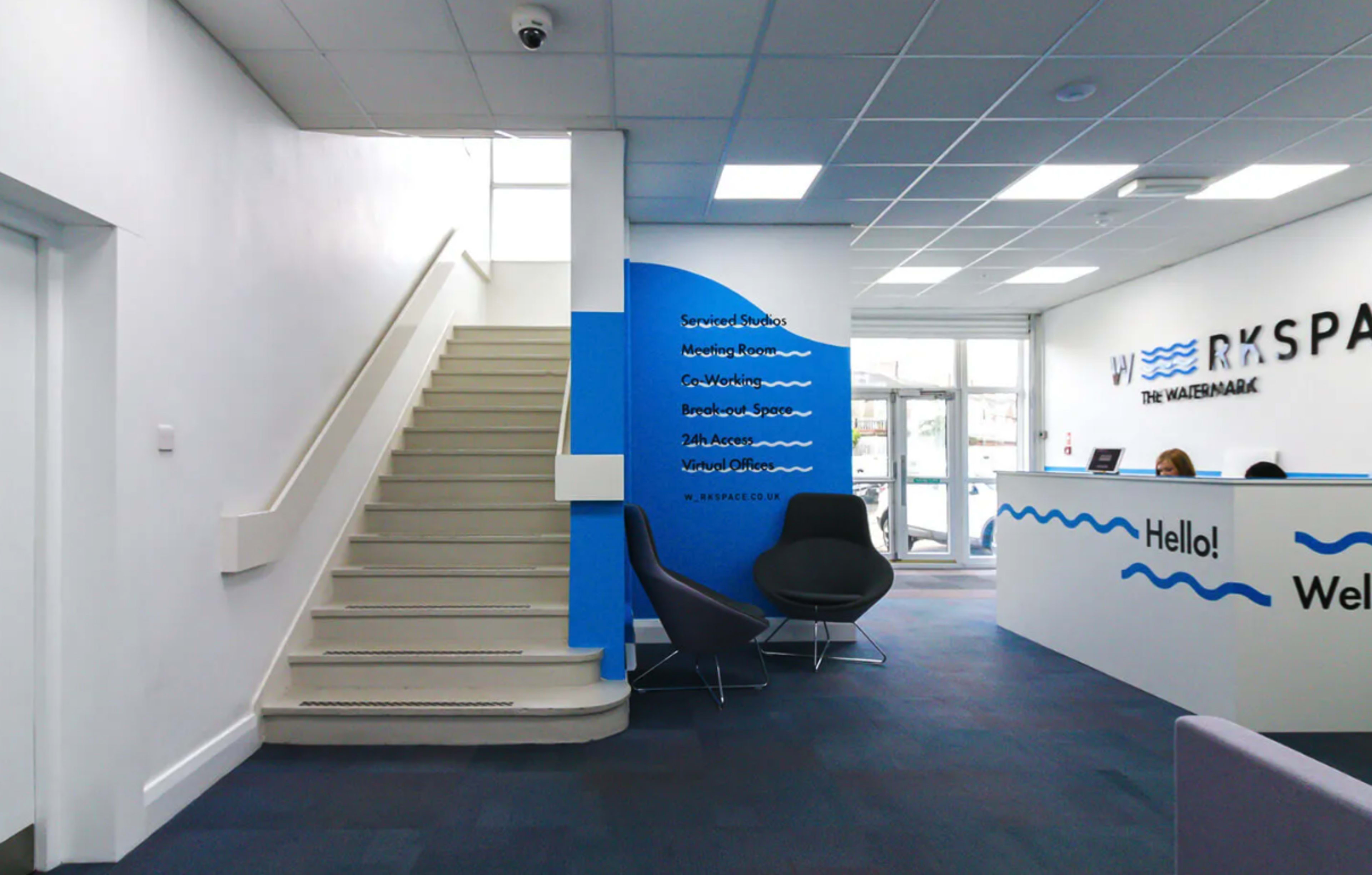 The image shows a modern office reception area with a blue accent wall, a seating area, and a staircase leading to an upper level.