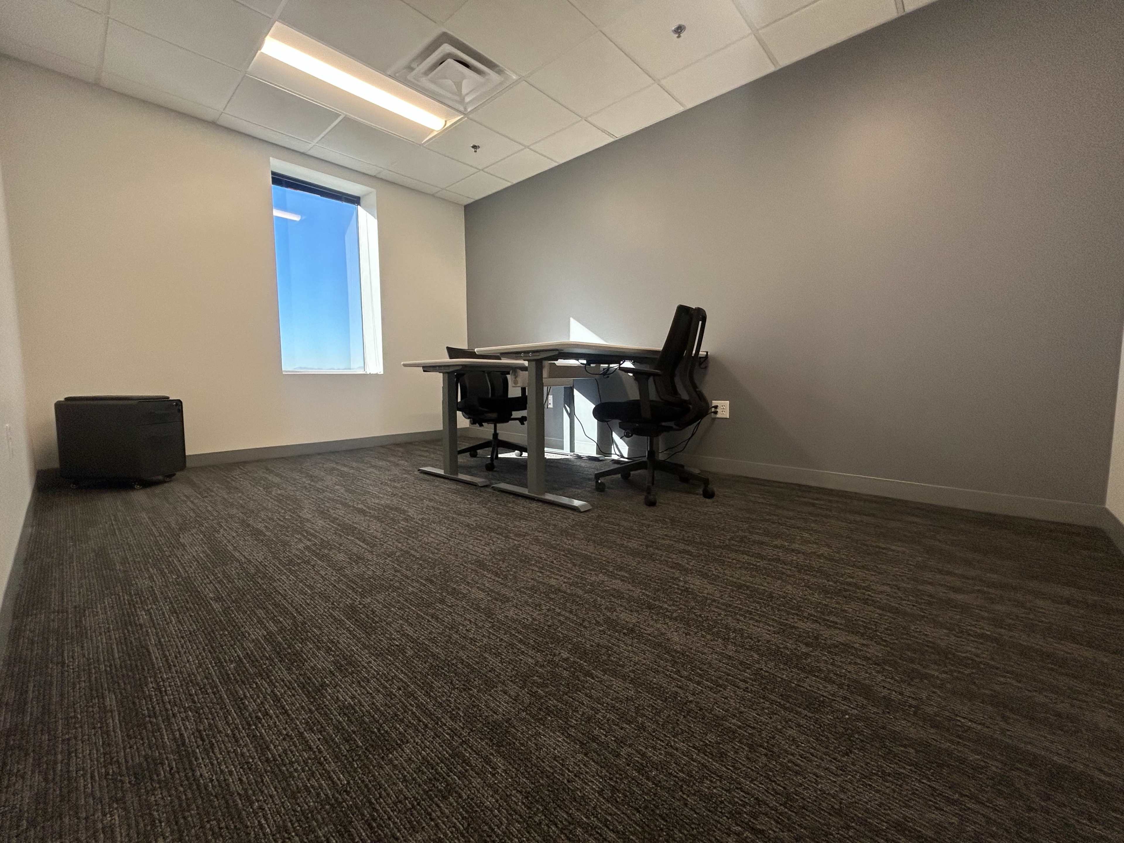 The image shows an empty office room with two chairs, a desk, a window providing natural light, and gray walls.