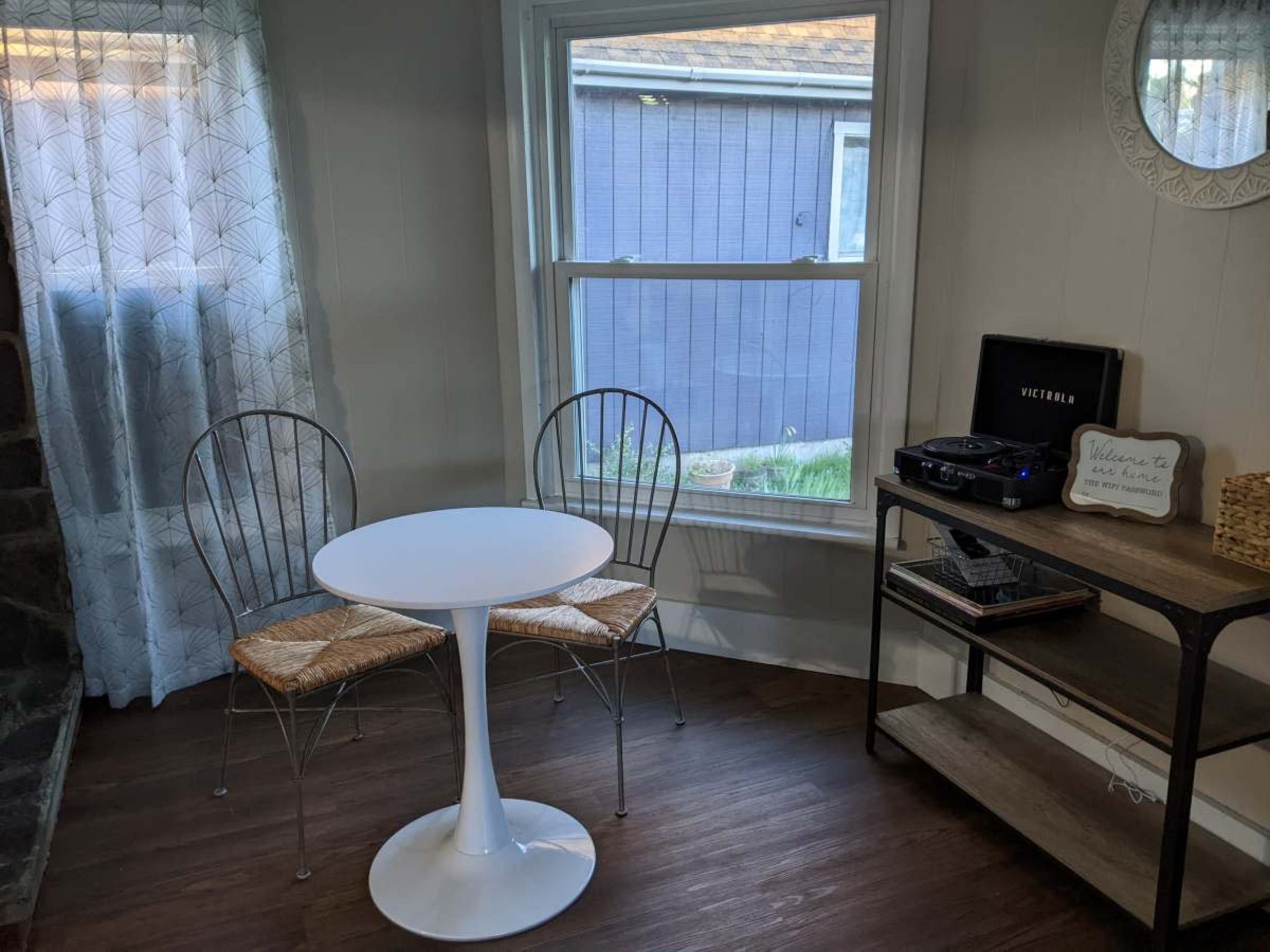 The image shows a small, minimalist dining area featuring a round white table with two wire chairs, a record player on a wooden shelf, and a window with sheer curtains.