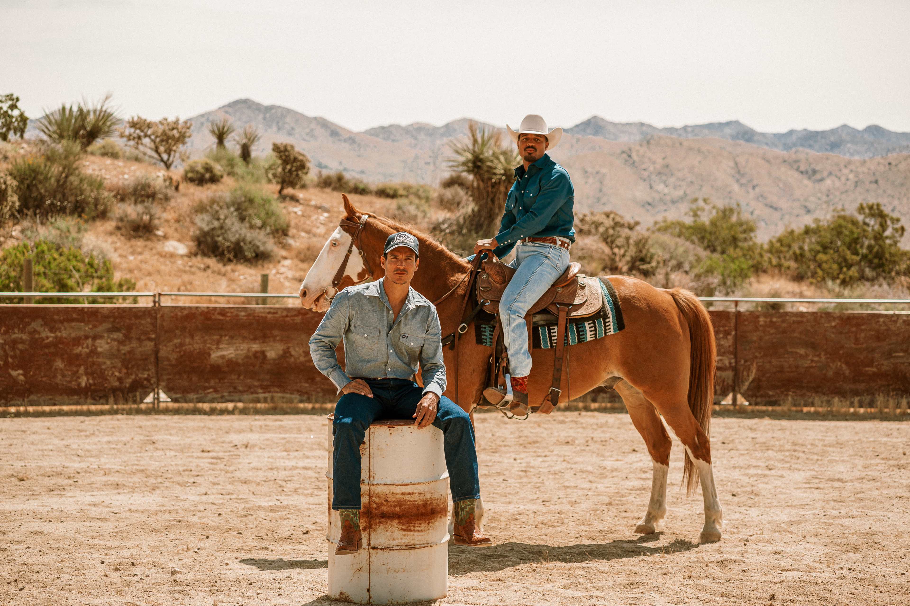 Two men pose outdoors near a horse in a sandy area with mountains in the background.