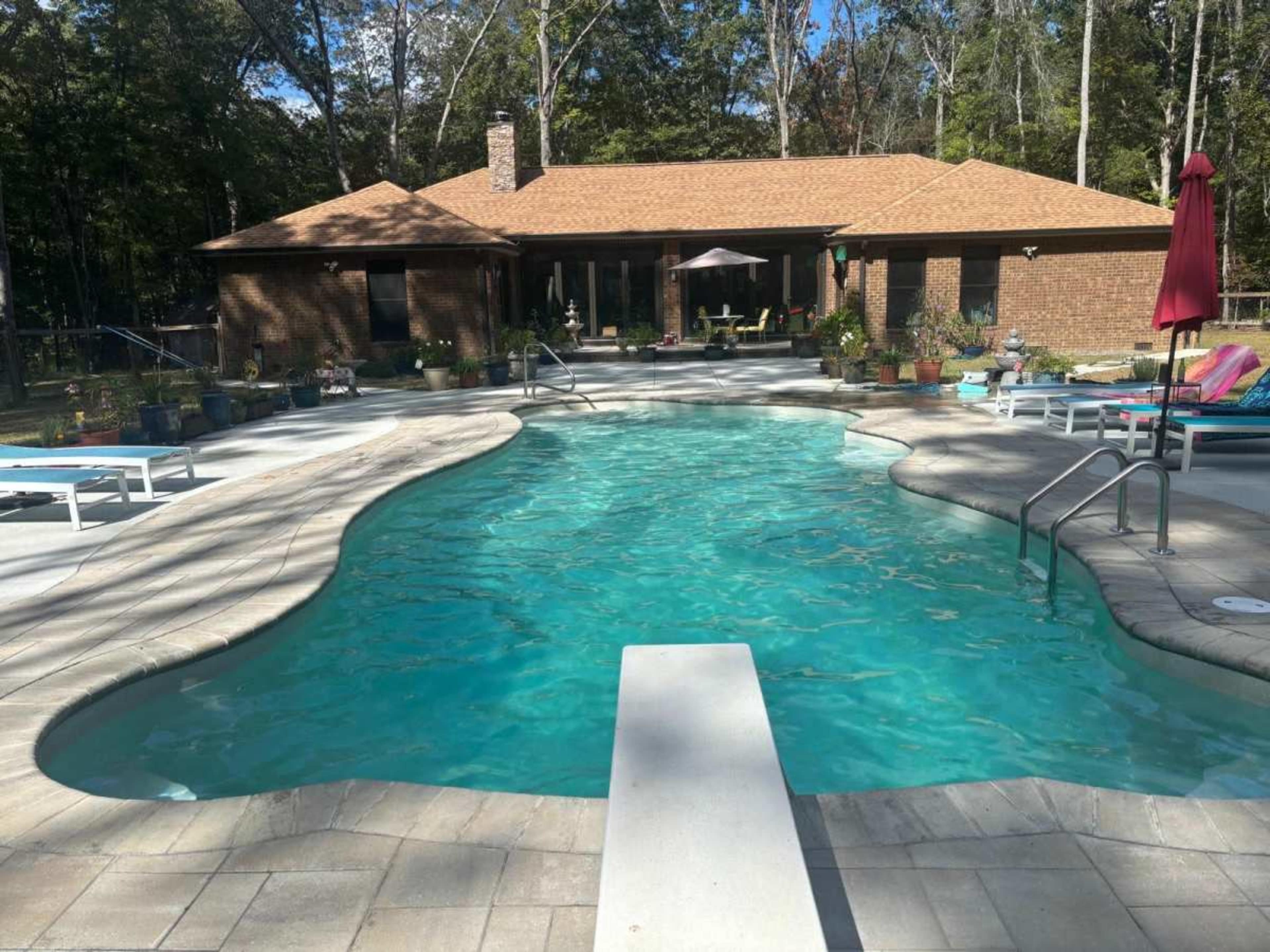 The image depicts a backyard swimming pool surrounded by a stone deck and a brick house, with lounge chairs and potted plants nearby.