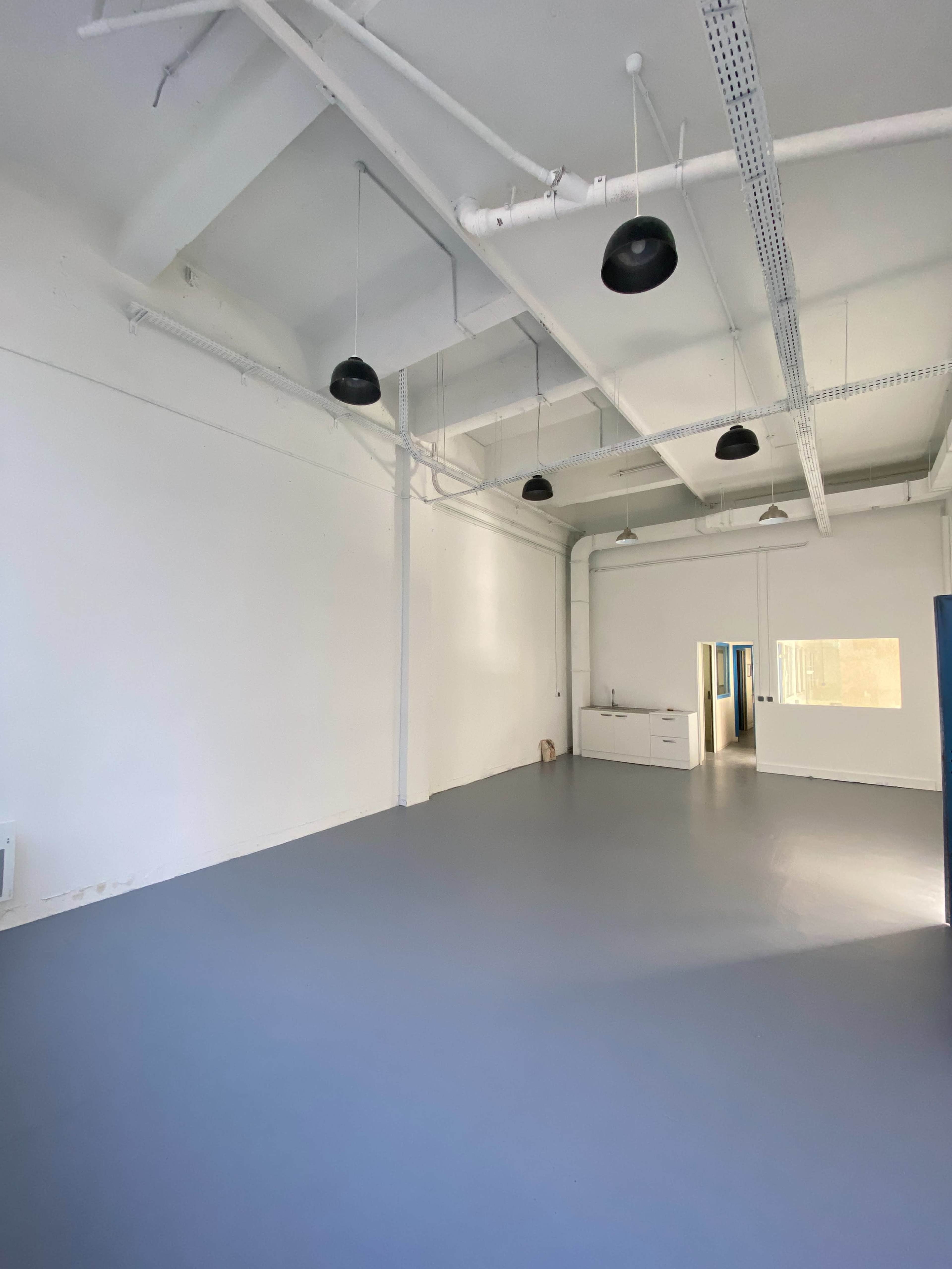 A spacious, empty room with white walls and gray flooring, featuring several black ceiling lights and a small kitchenette in the background.