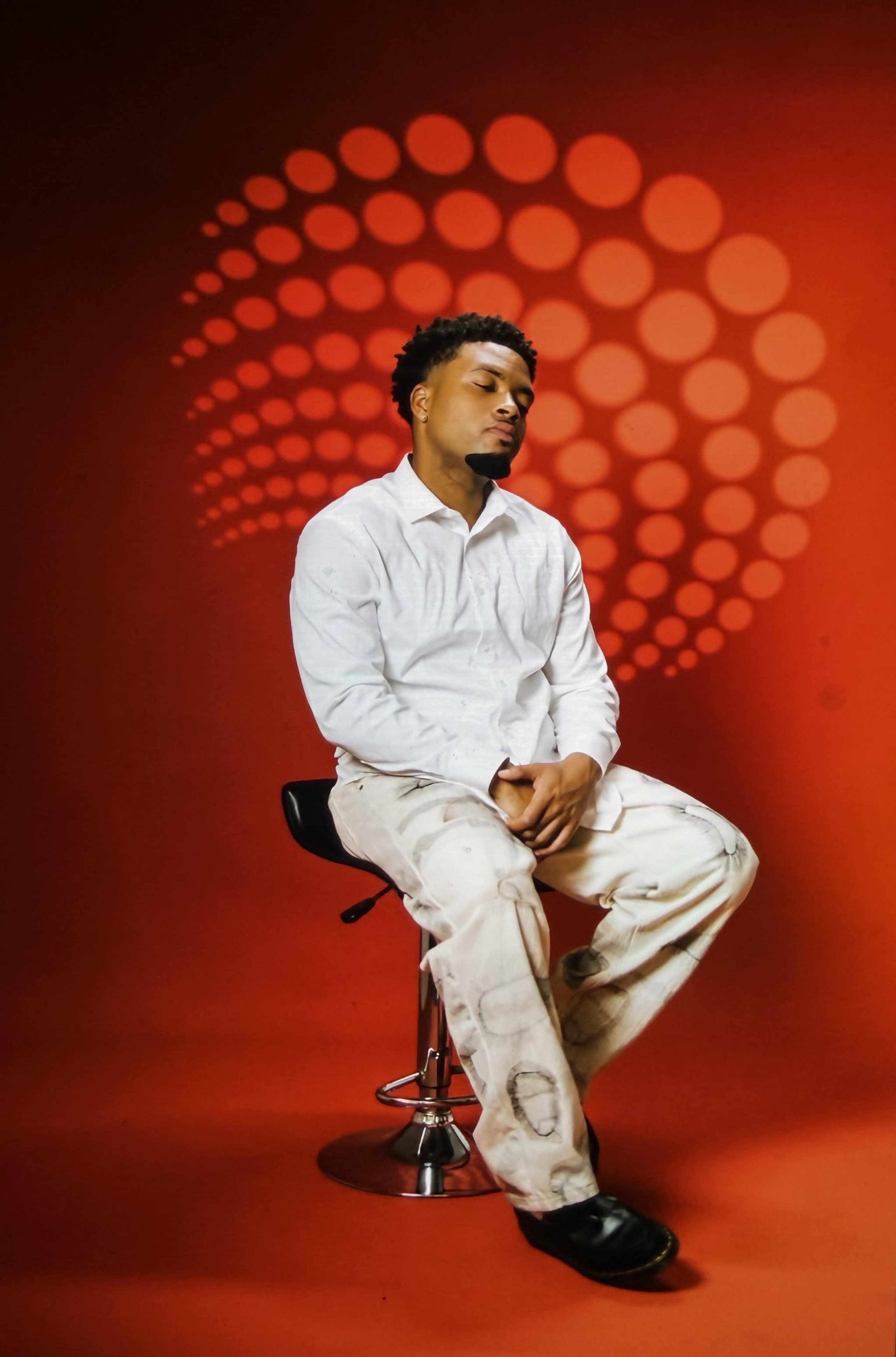 A person sits on a stool against a vivid red backdrop with circular shadows.