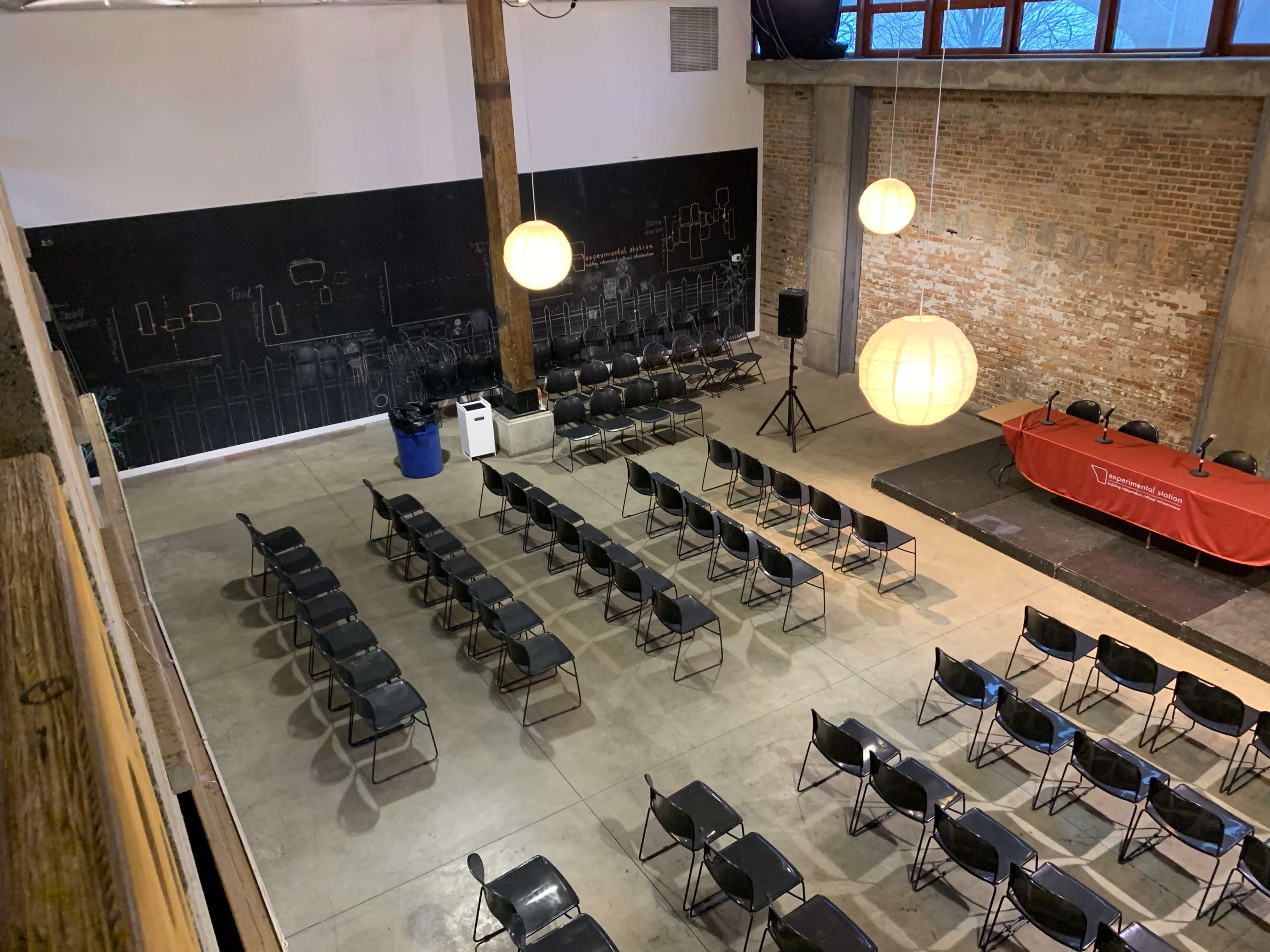 A spacious event room with rows of black chairs, a red tablecloth on a stage, and large pendant lights hanging overhead.