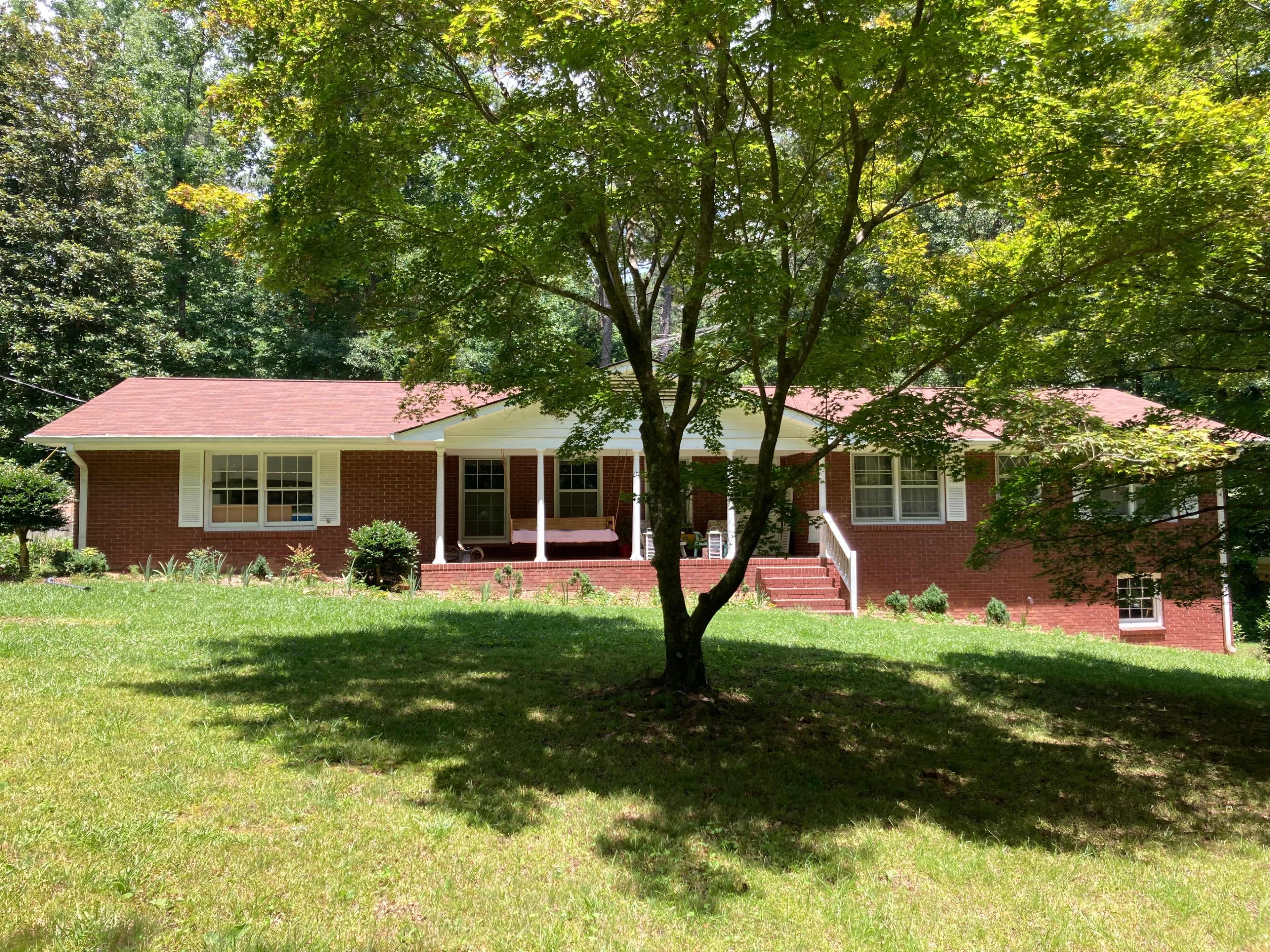 A red brick ranch-style house with a front porch is set on a grassy slope and surrounded by trees.