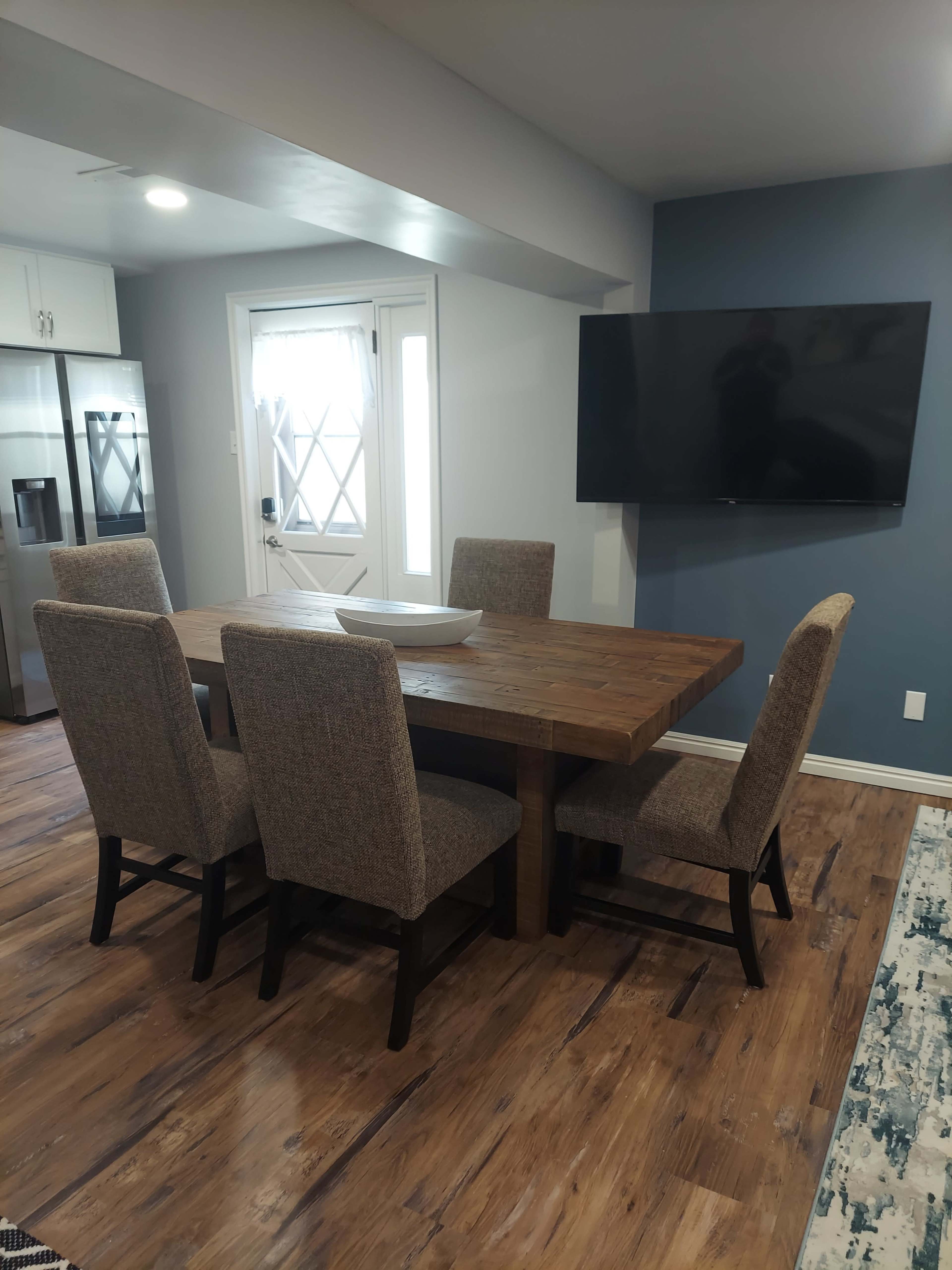 A dining area with a wooden table surrounded by four chairs, a wall-mounted television, and a door leading outside.