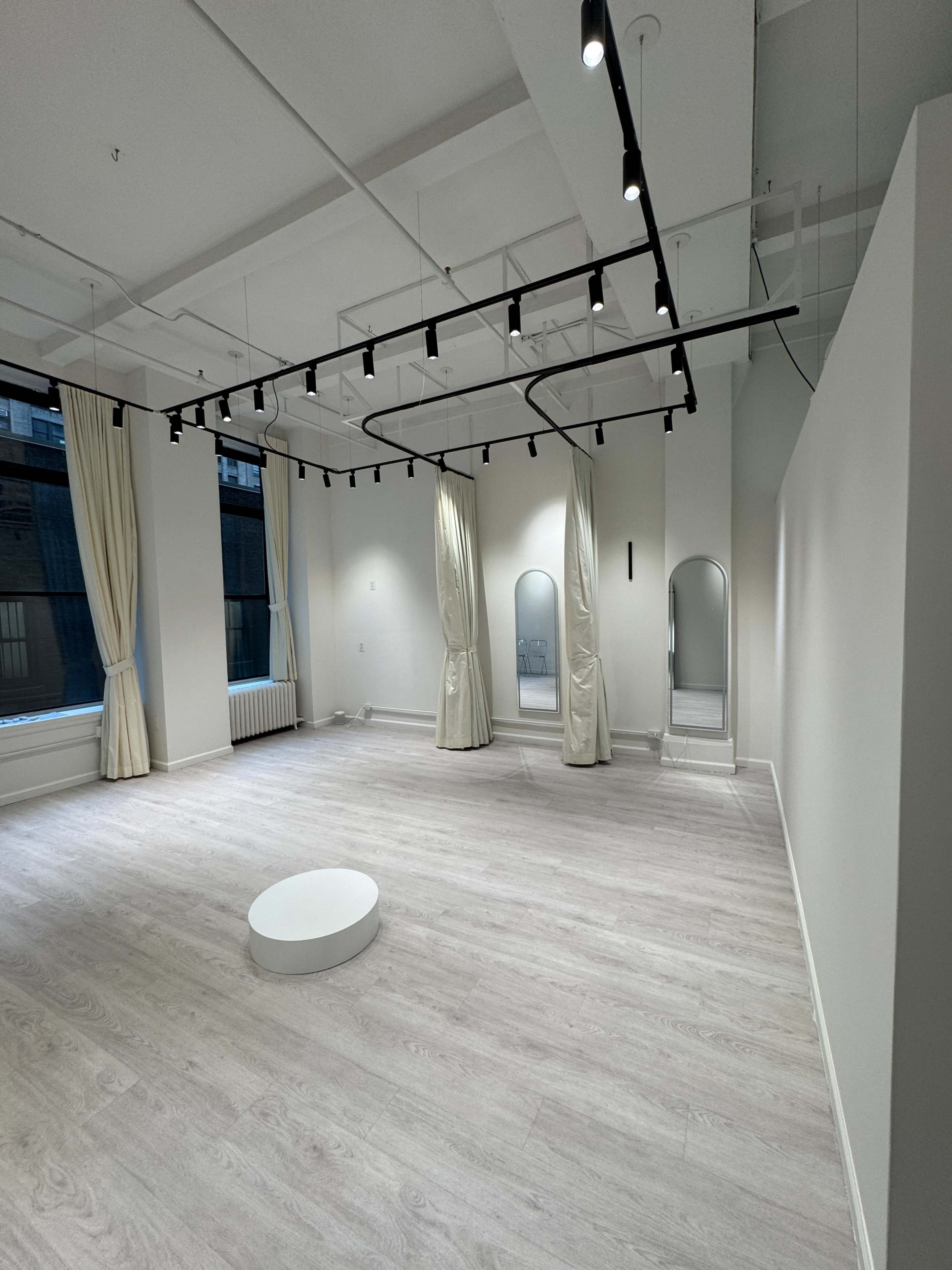 The image shows a minimalist, empty studio room with light wooden floors, white walls, large windows, and three floor-length mirrors.