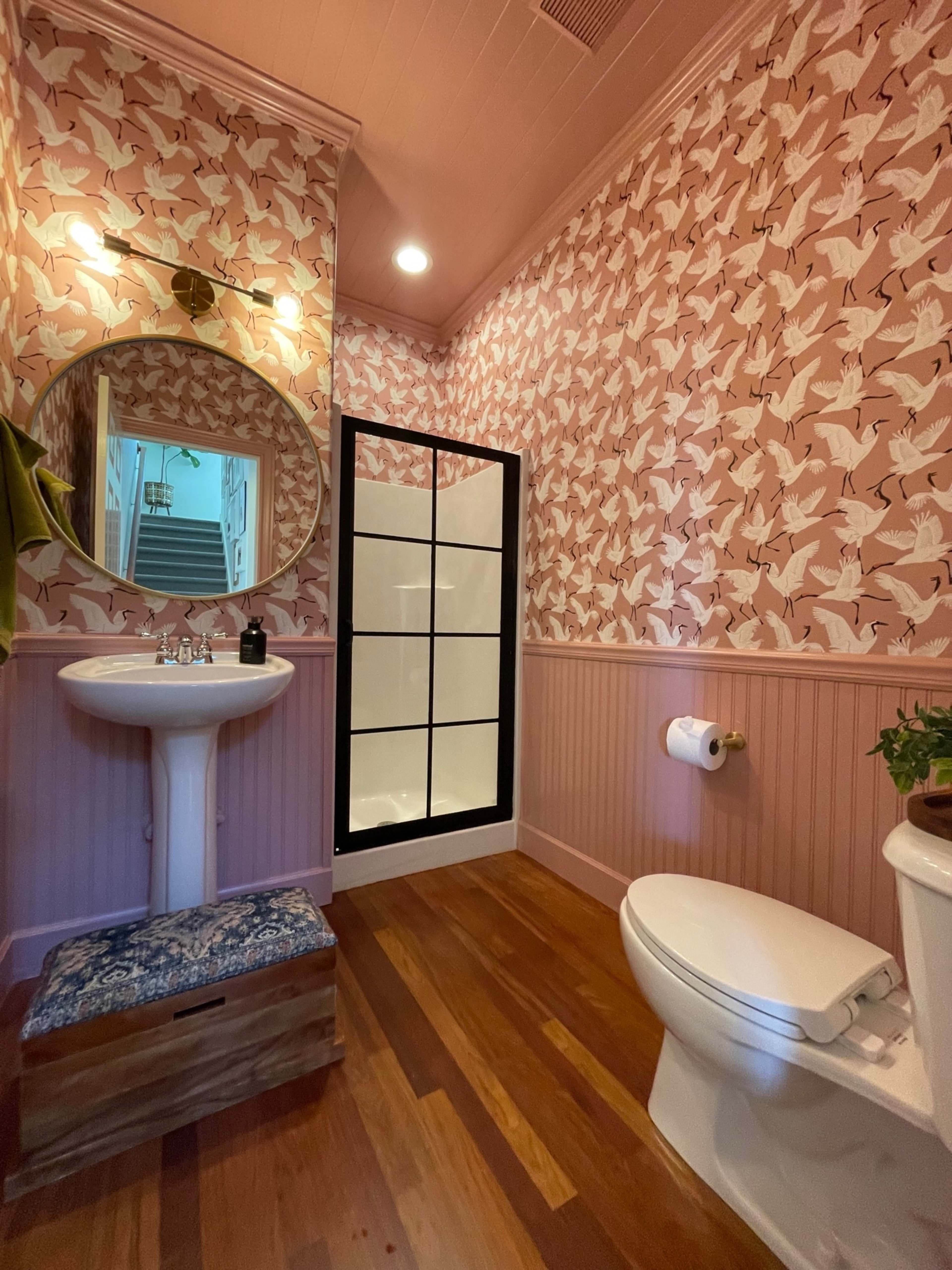 A small bathroom features a pink patterned wallpaper, a circular mirror above a pedestal sink, a shower with a glass door, a toilet, and wooden flooring.