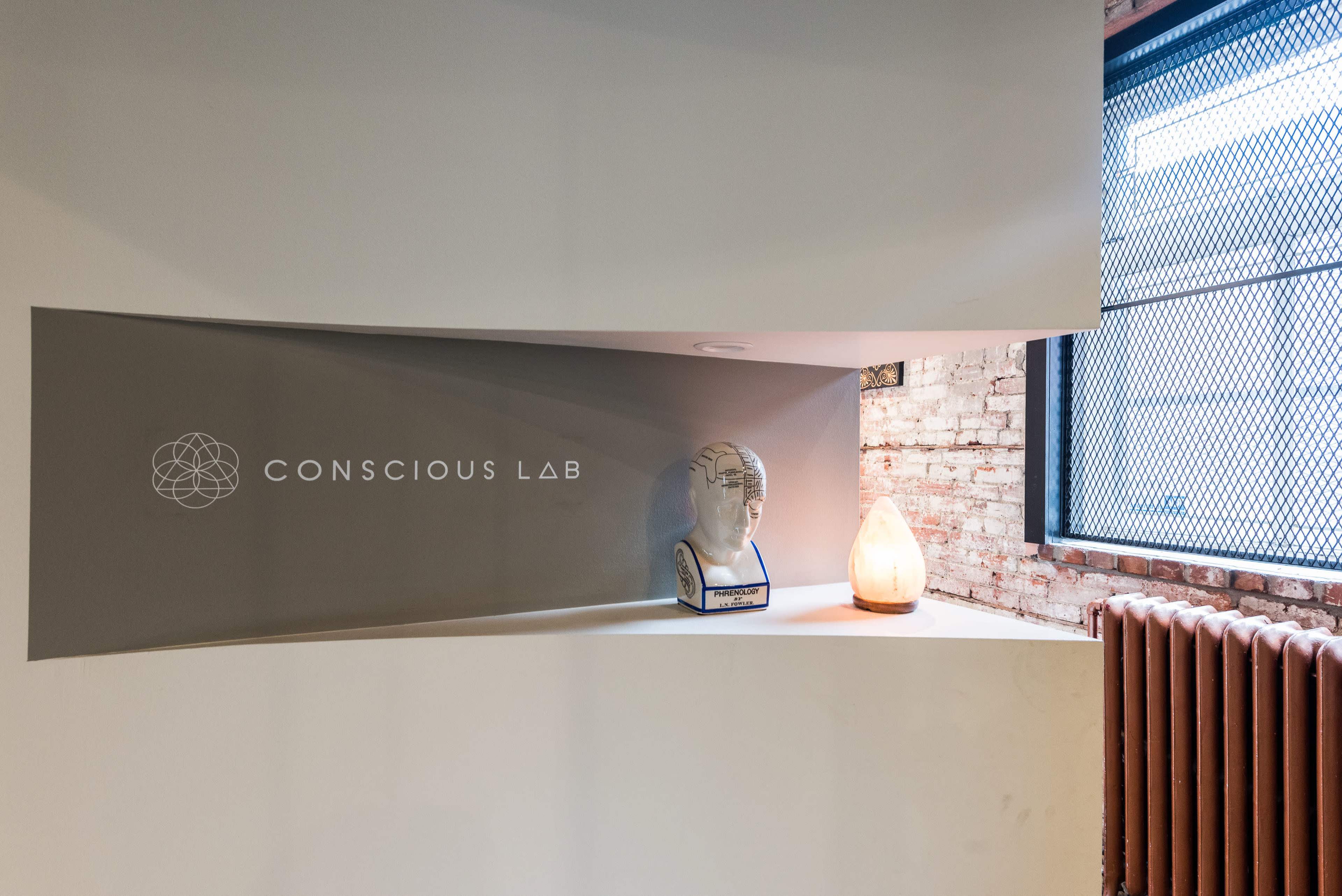 A modern interior space featuring a wall with the logo "Conscious Lab," a decorative bust, and a soft-lit lamp, alongside a brick wall and a window with a metal grid.