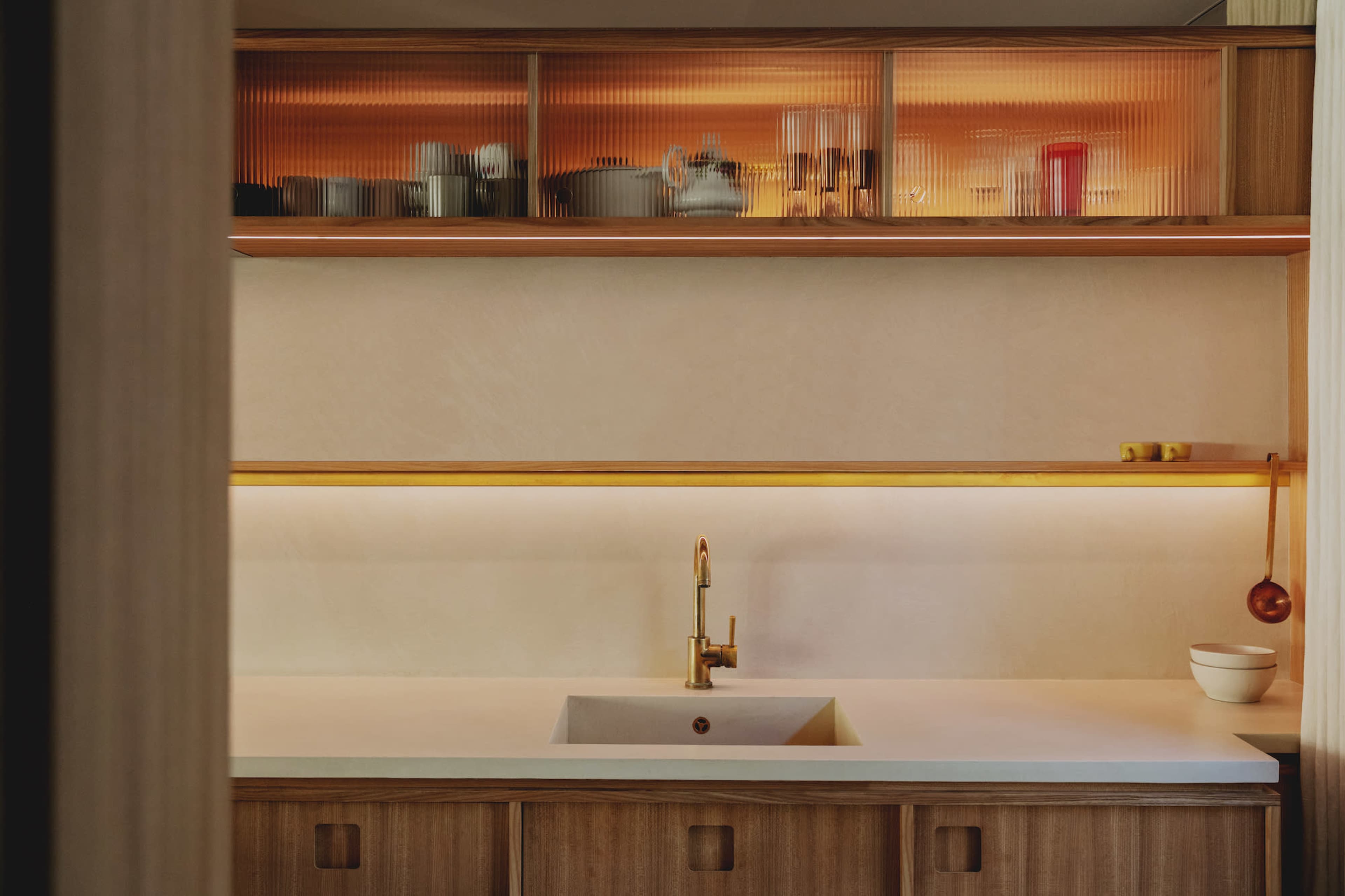 The image shows a modern kitchen with a sleek countertop, a gold faucet, and illuminated shelves displaying various kitchen items.