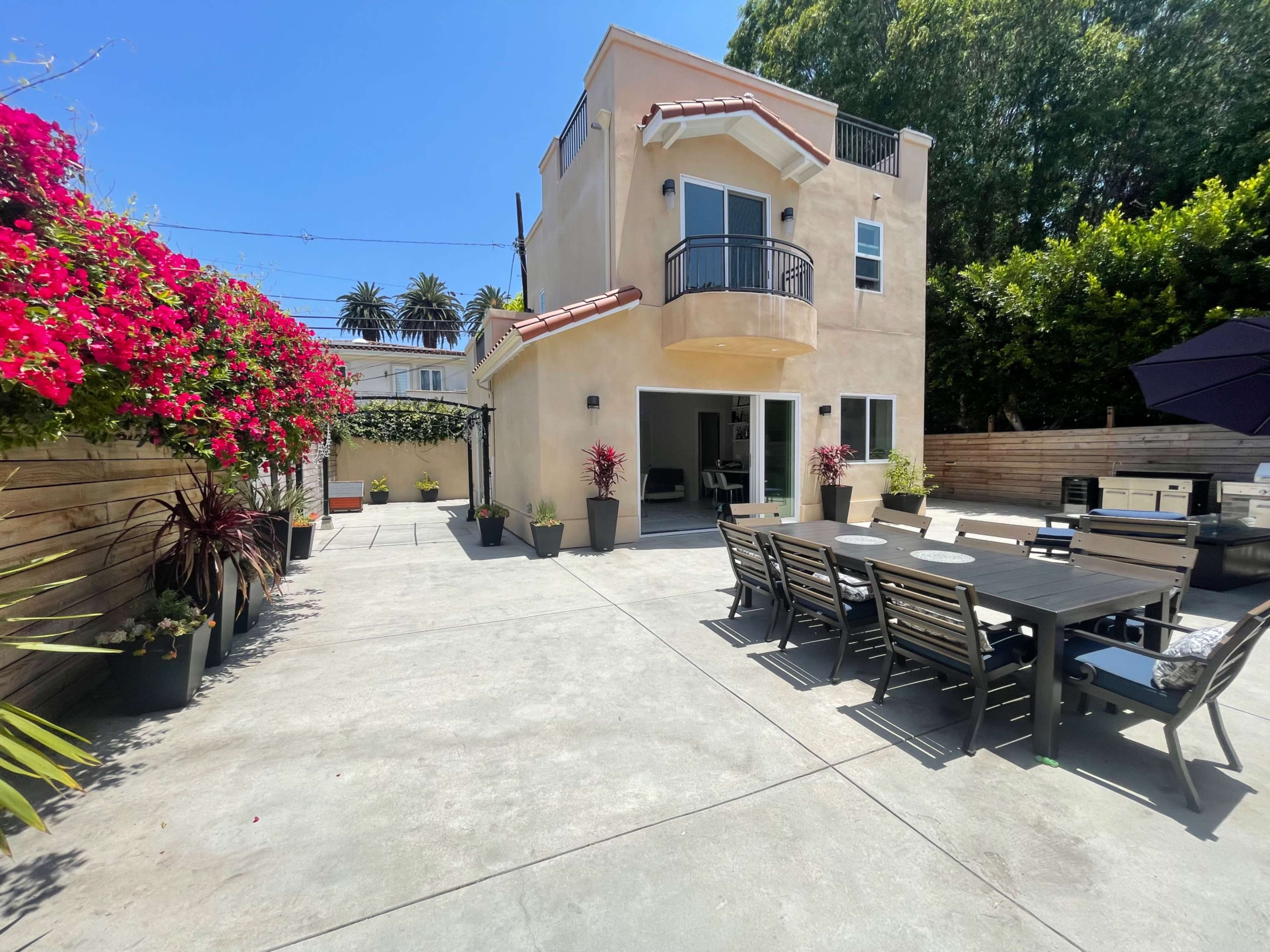 The image shows a spacious outdoor patio area with a dining table, surrounded by potted plants and flowering bushes, adjacent to a two-story beige house.