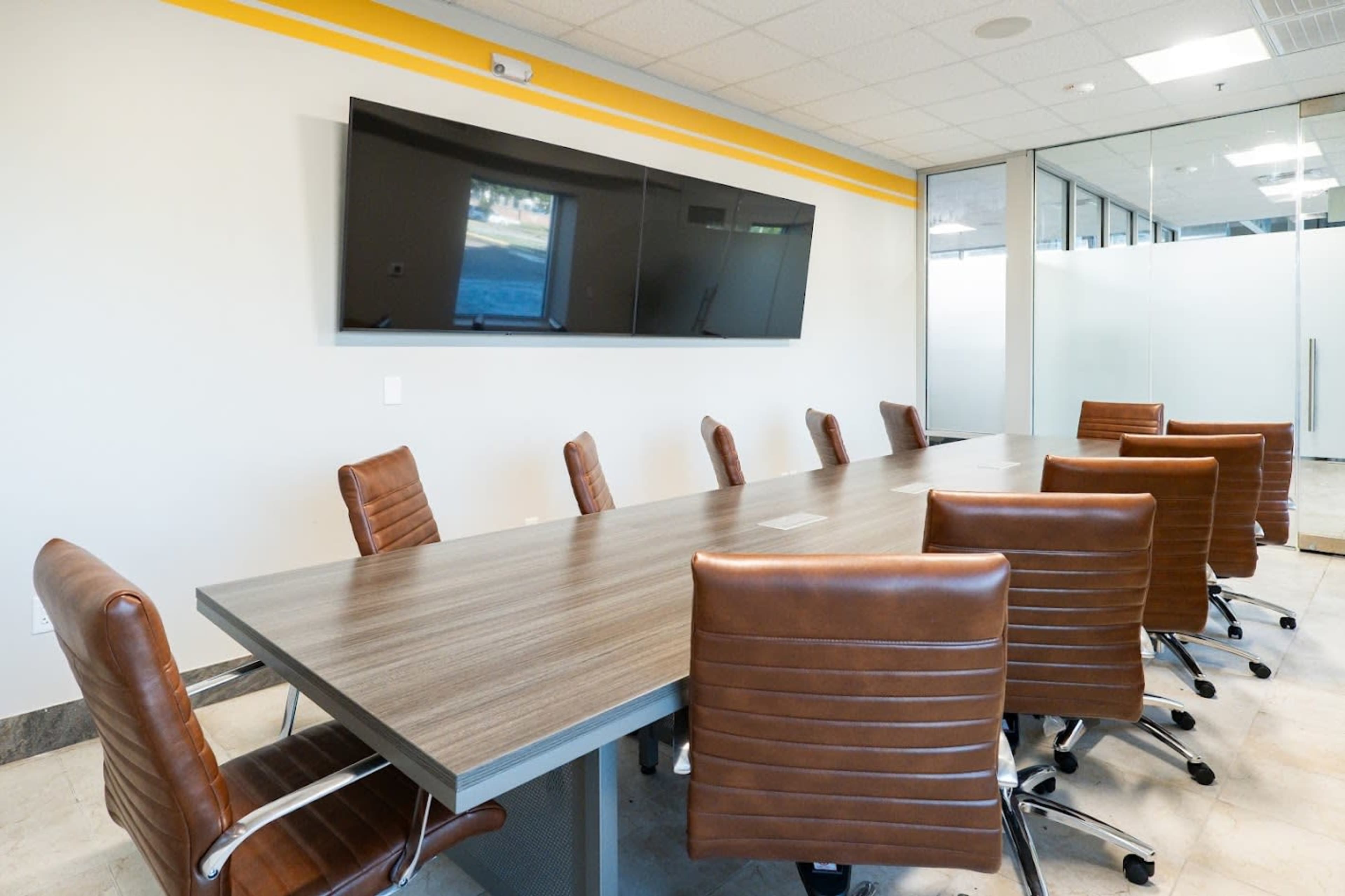 The image shows a modern conference room with a long wooden table surrounded by eight brown leather chairs and a large flat-screen television mounted on the wall.