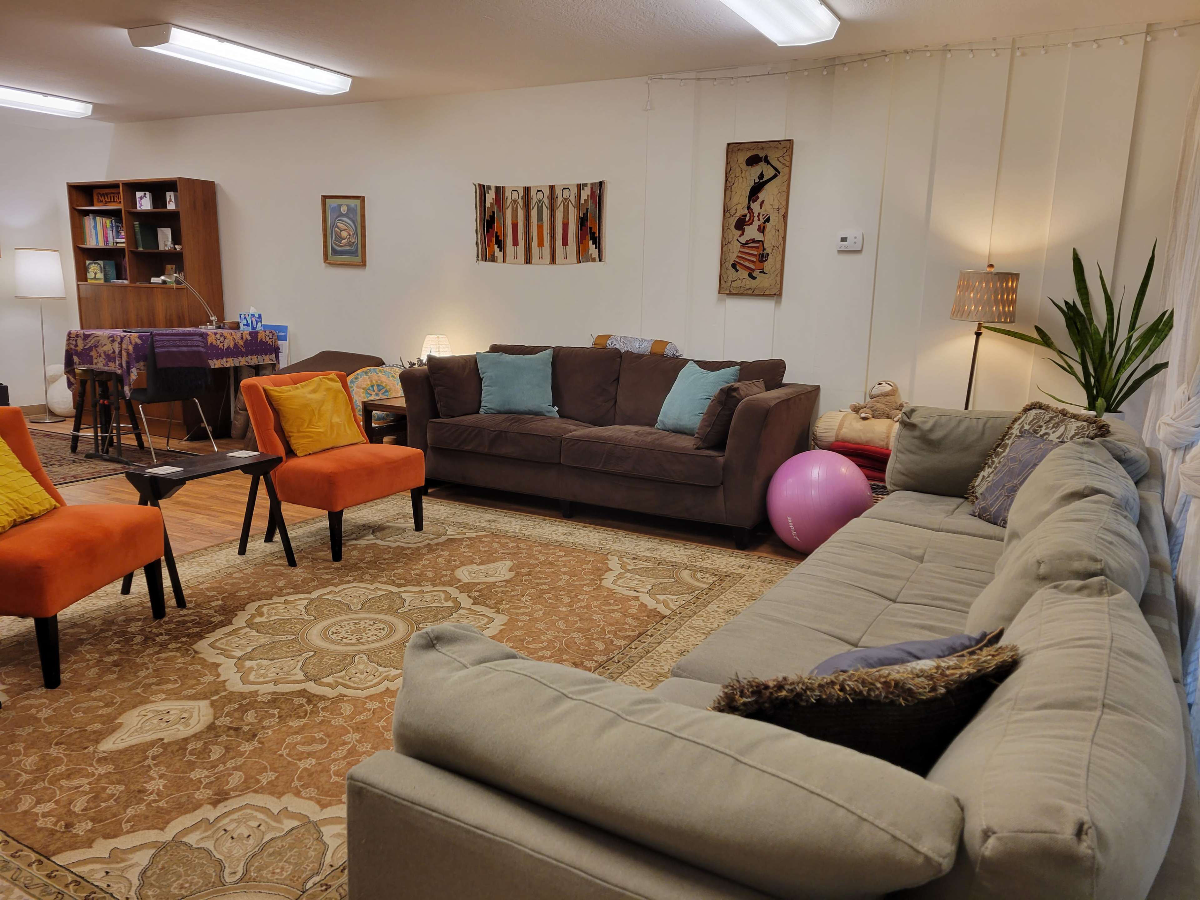 A cozy room is arranged with a large sectional sofa, orange armchairs, a coffee table, and a rug, featuring decorative art on the walls and plants in the corners.