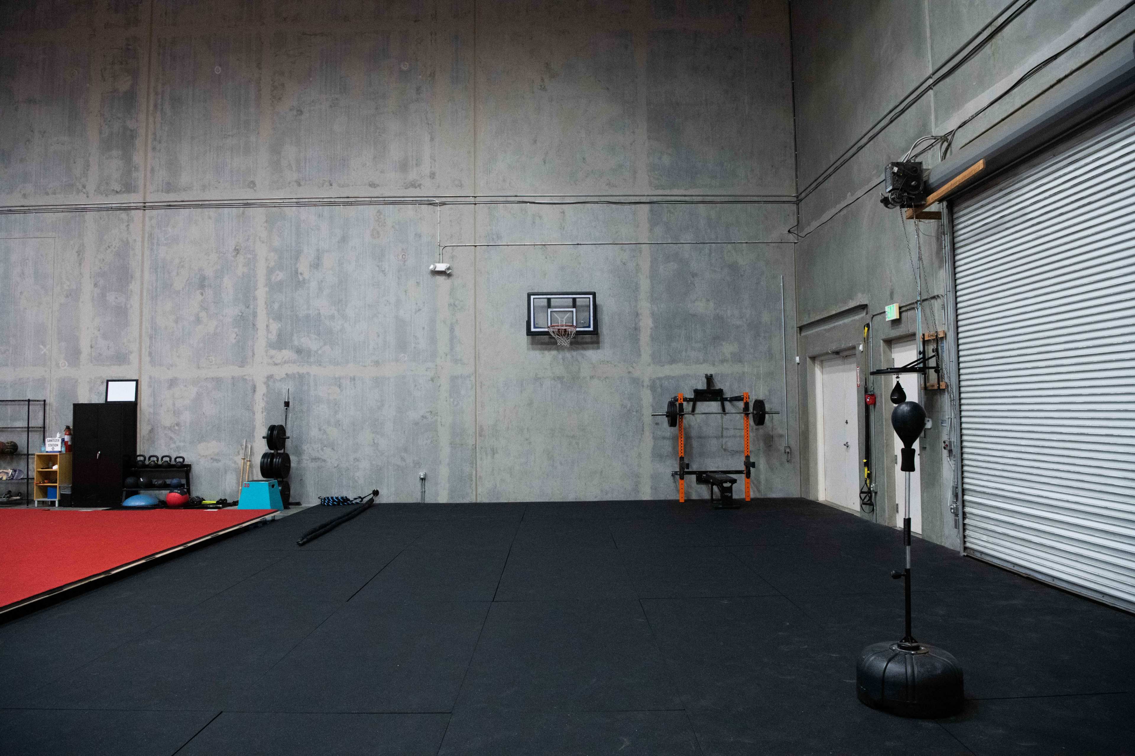 A spacious gym with a gray concrete wall, a basketball hoop, a punching bag on a stand, and exercise equipment along one side.