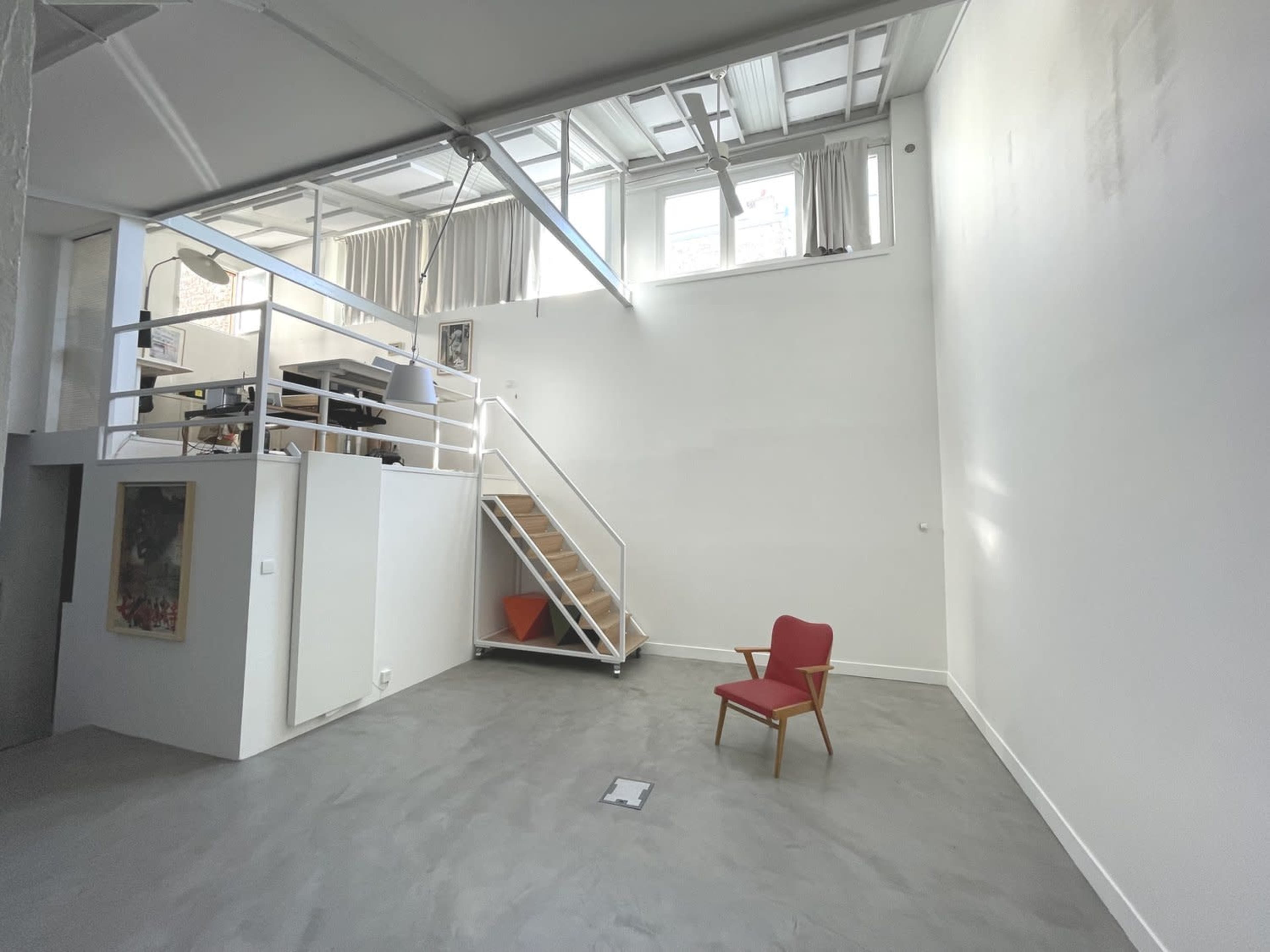 A spacious room with a wooden staircase leading to a loft area, a red chair on the floor, and large windows along the upper portion of the wall.