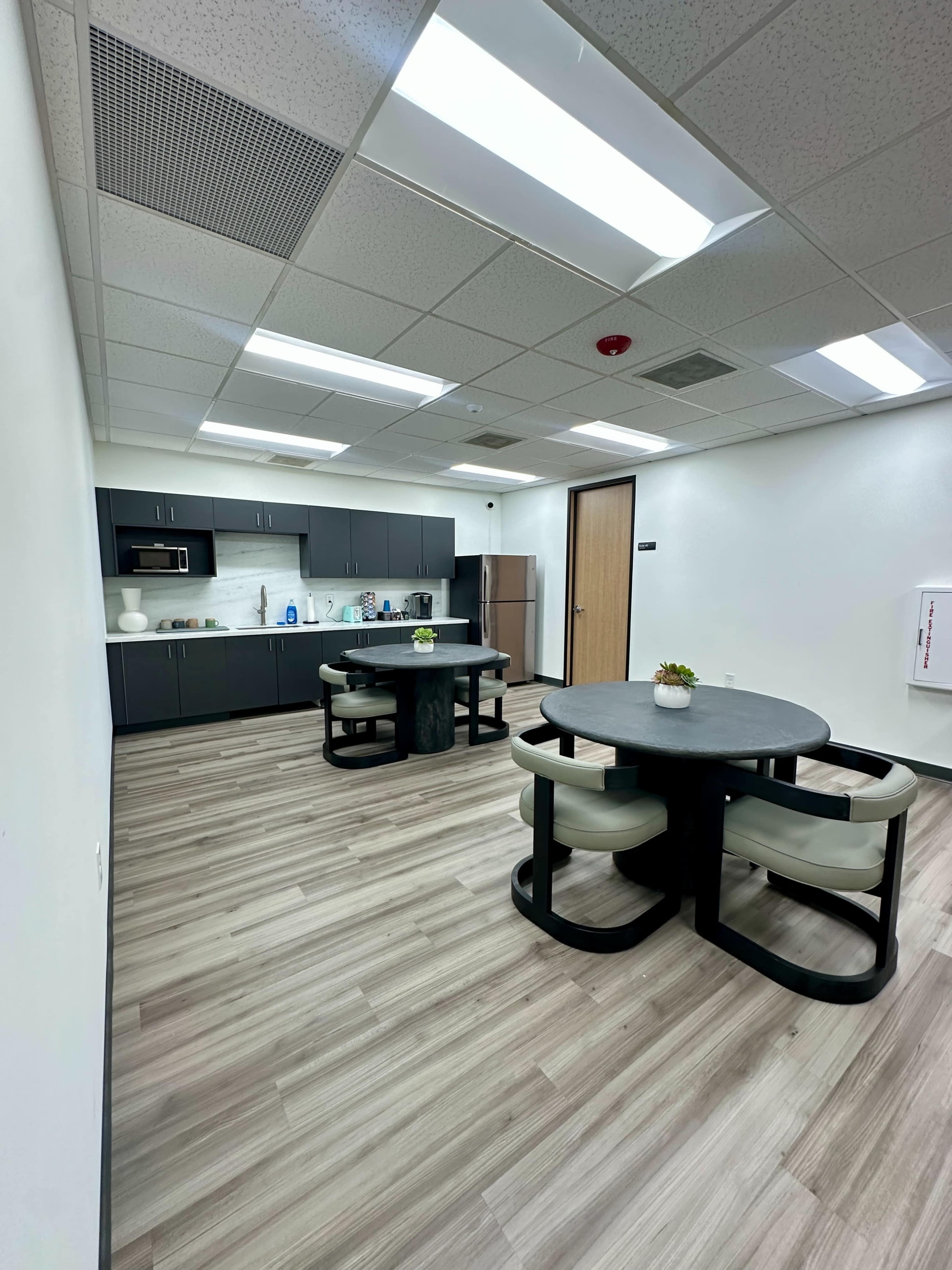 The image shows a modern break room with two round tables and a kitchen area featuring dark cabinets and stainless steel appliances.