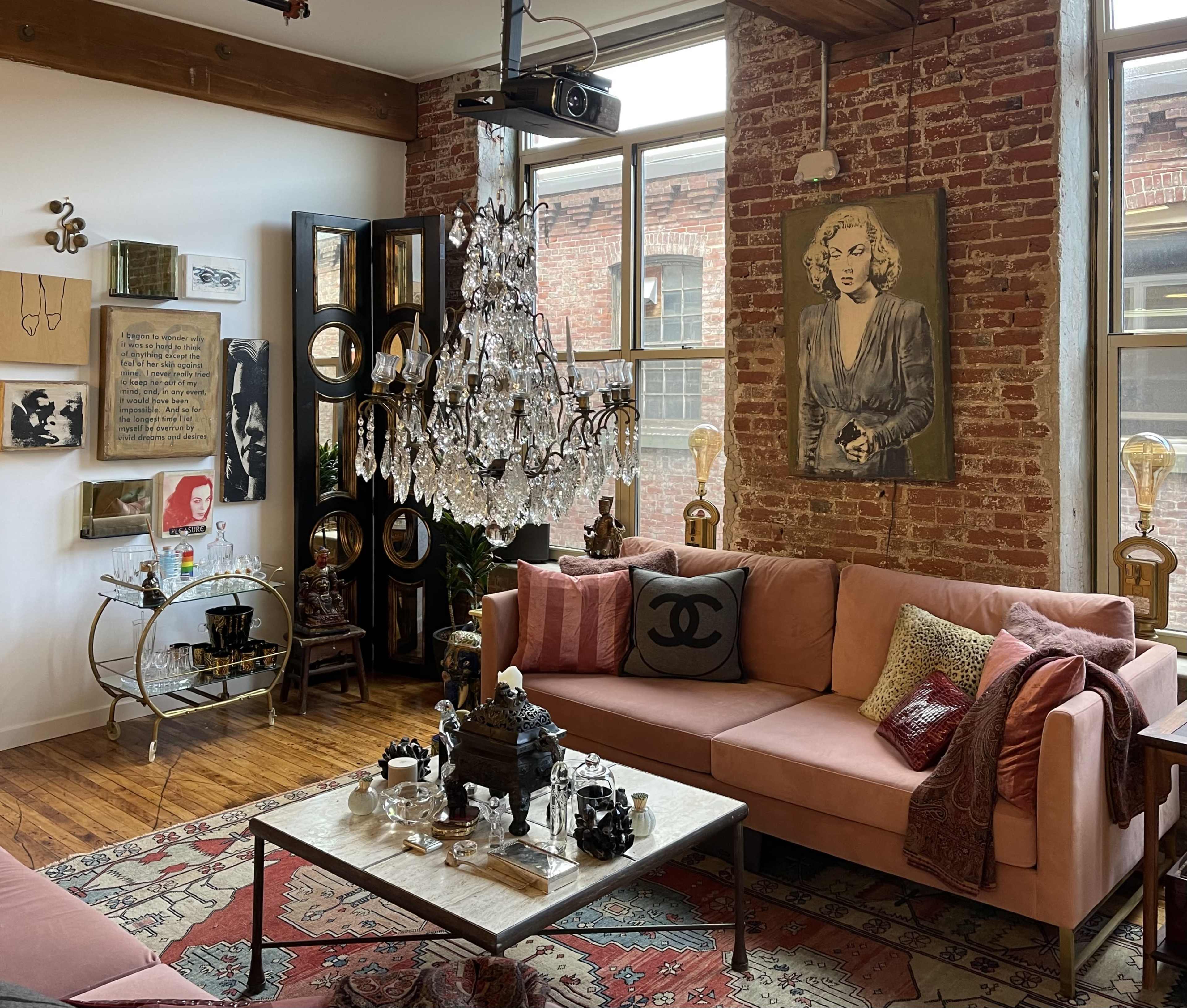 The image features a stylish living room with exposed brick walls, a pink sectional sofa, a crystal chandelier, and various decorative items.