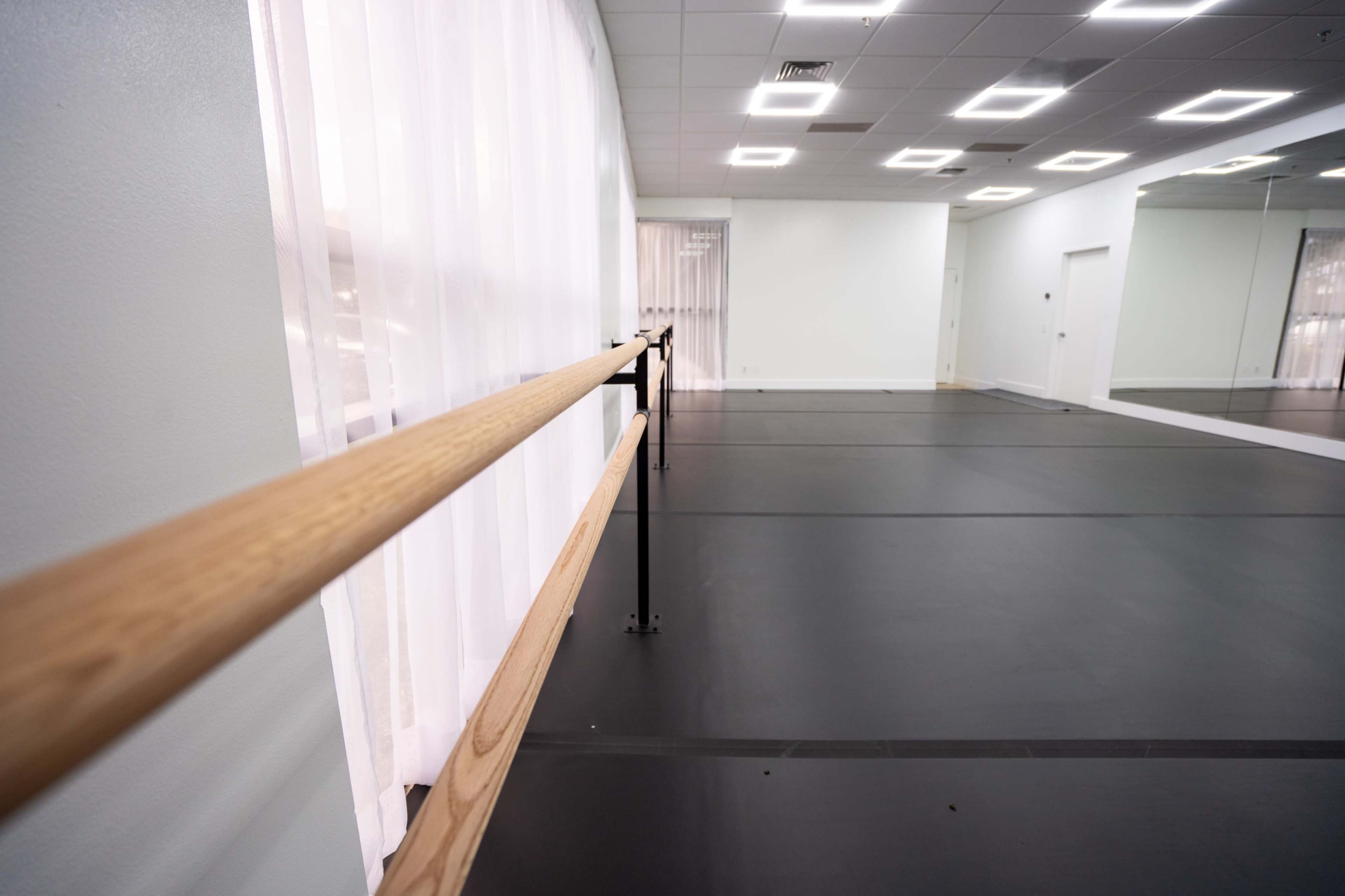 The image shows a dance studio with a wooden ballet bar, large windows covered with sheer curtains, and mirrored walls.