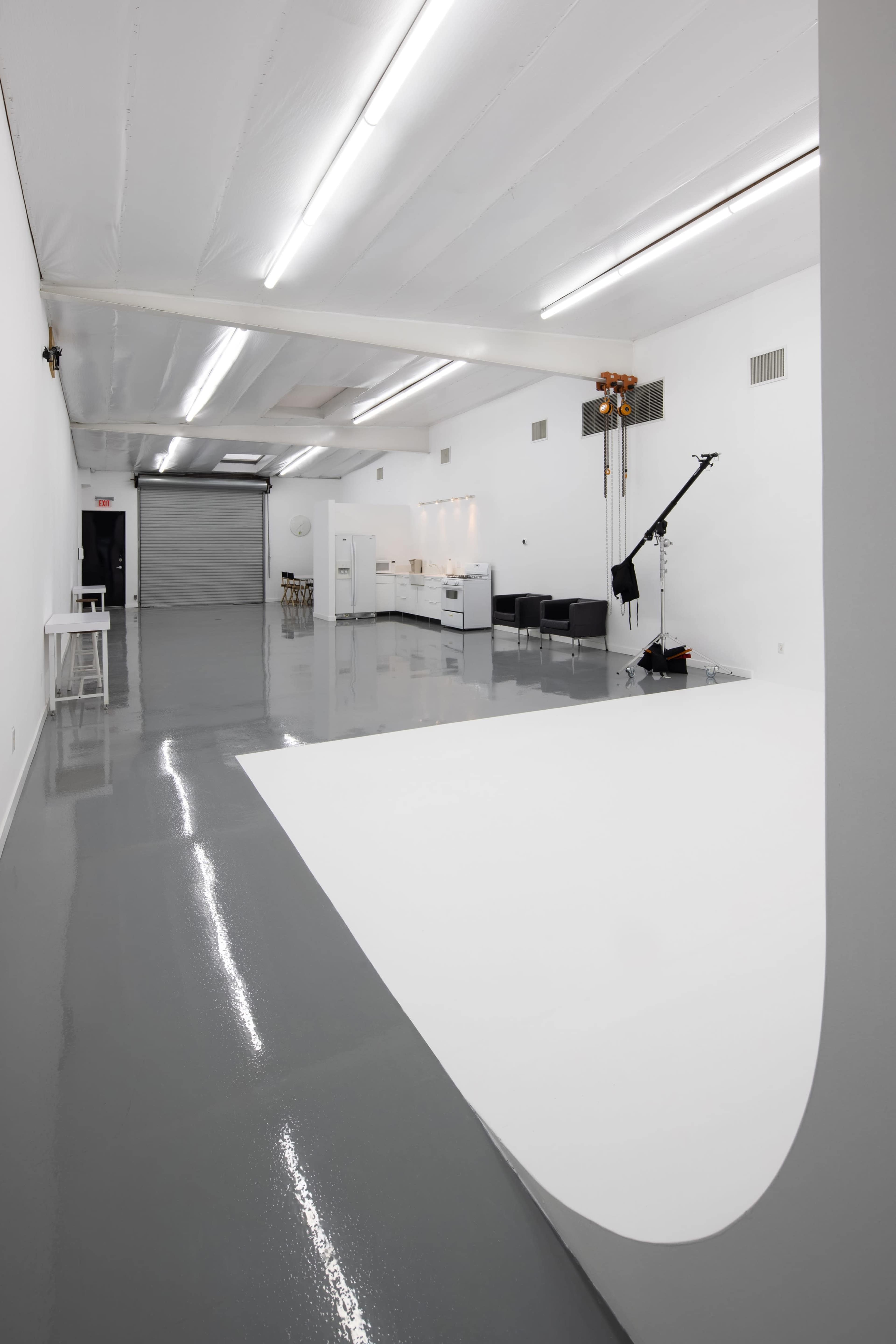 The image shows a spacious, empty studio with a gray floor, a kitchenette area, and a backdrop setup in the foreground.