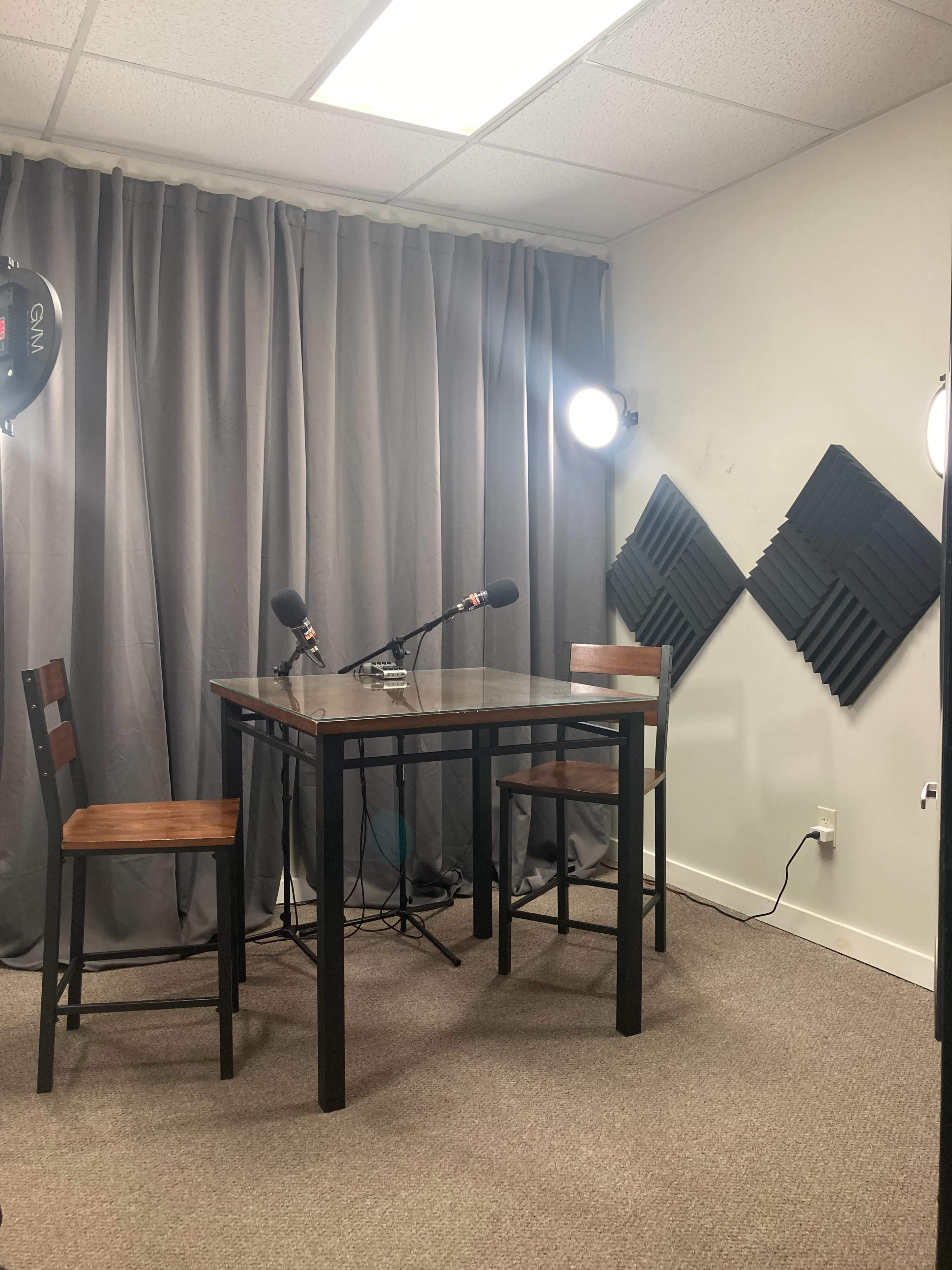 A small recording setup with a table and two chairs, framed by gray curtains and equipped with microphones and softbox lights.