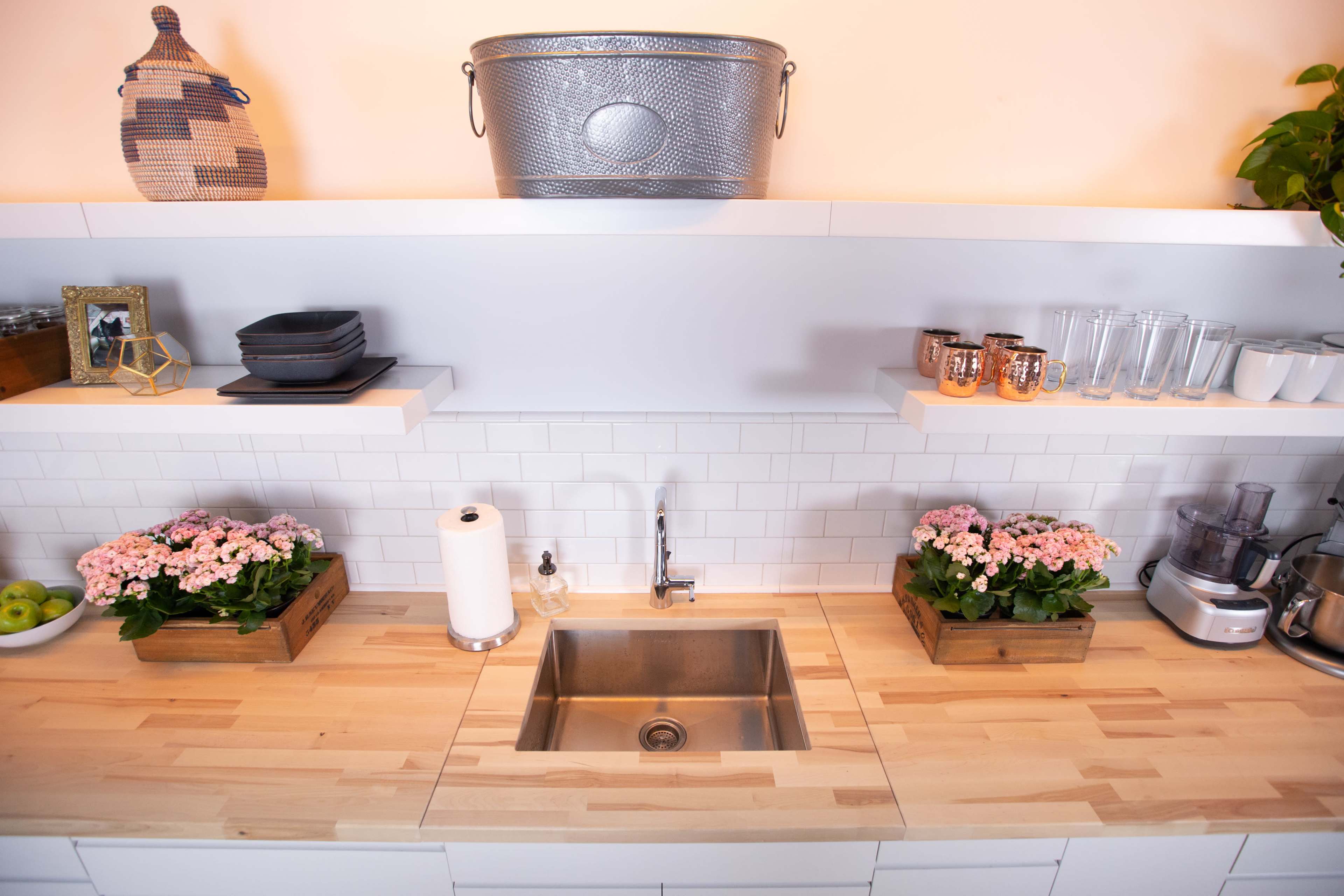 The image shows a modern kitchen sink area with a wooden countertop, white tiled backsplash, and decorative potted flowers on either side.