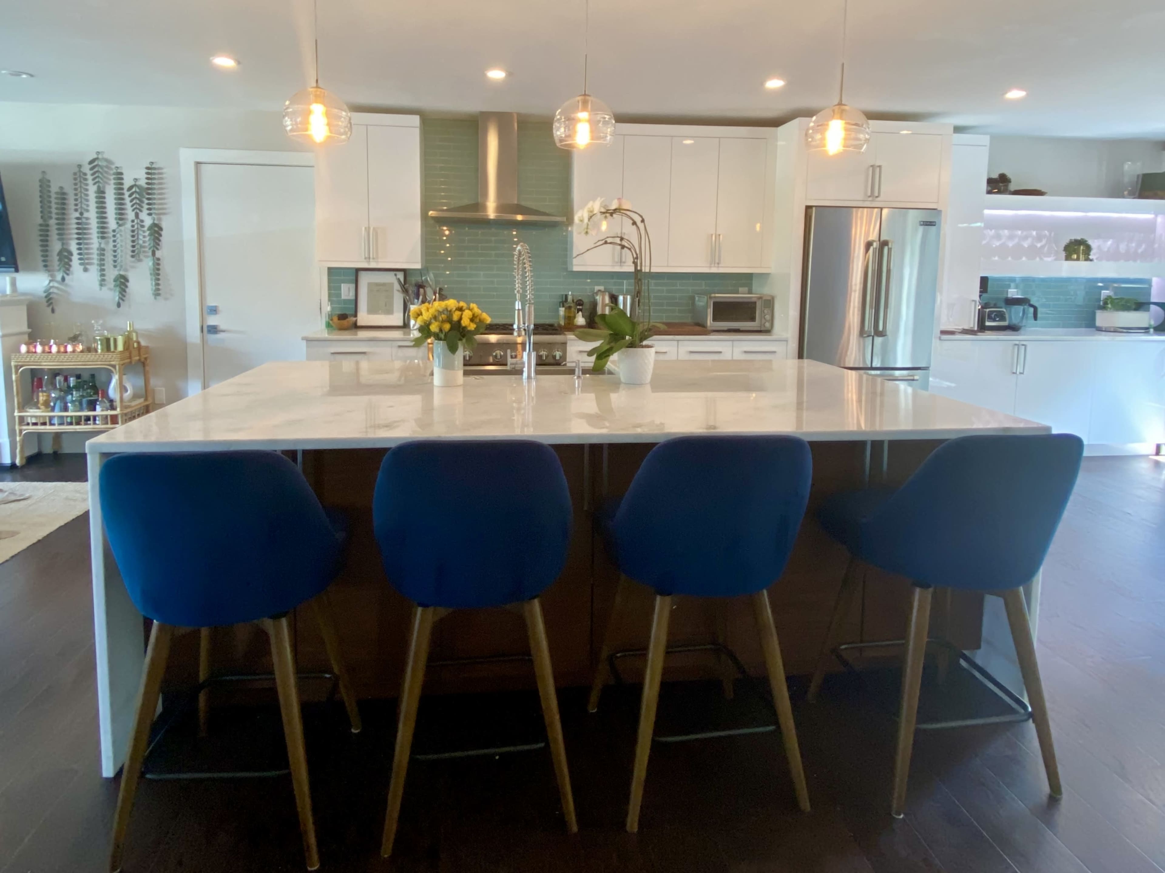 A modern kitchen features a large marble island with blue bar stools and stainless steel appliances.