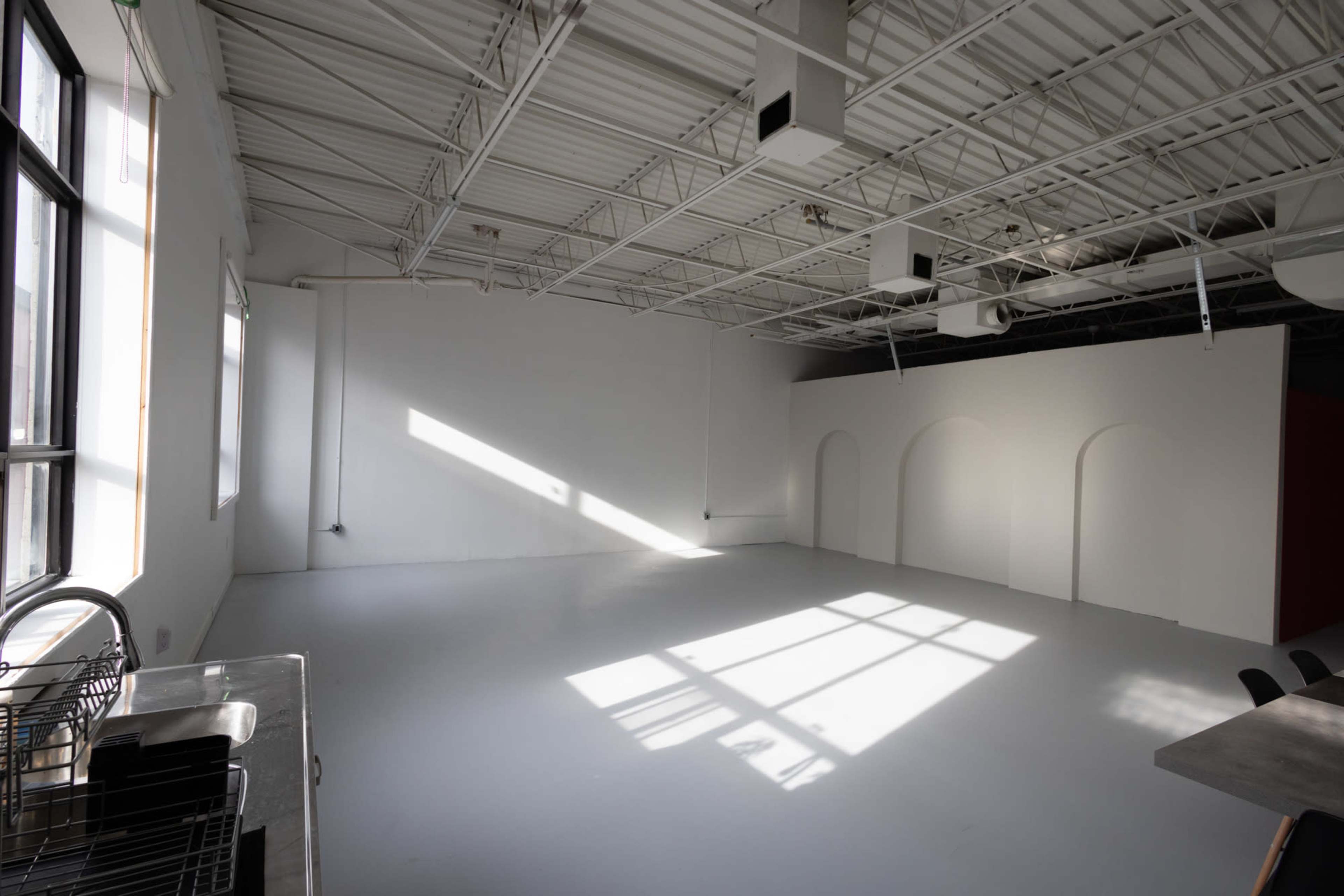 The image shows a spacious, well-lit studio with large windows and a white floor, featuring minimalistic archways on one wall.