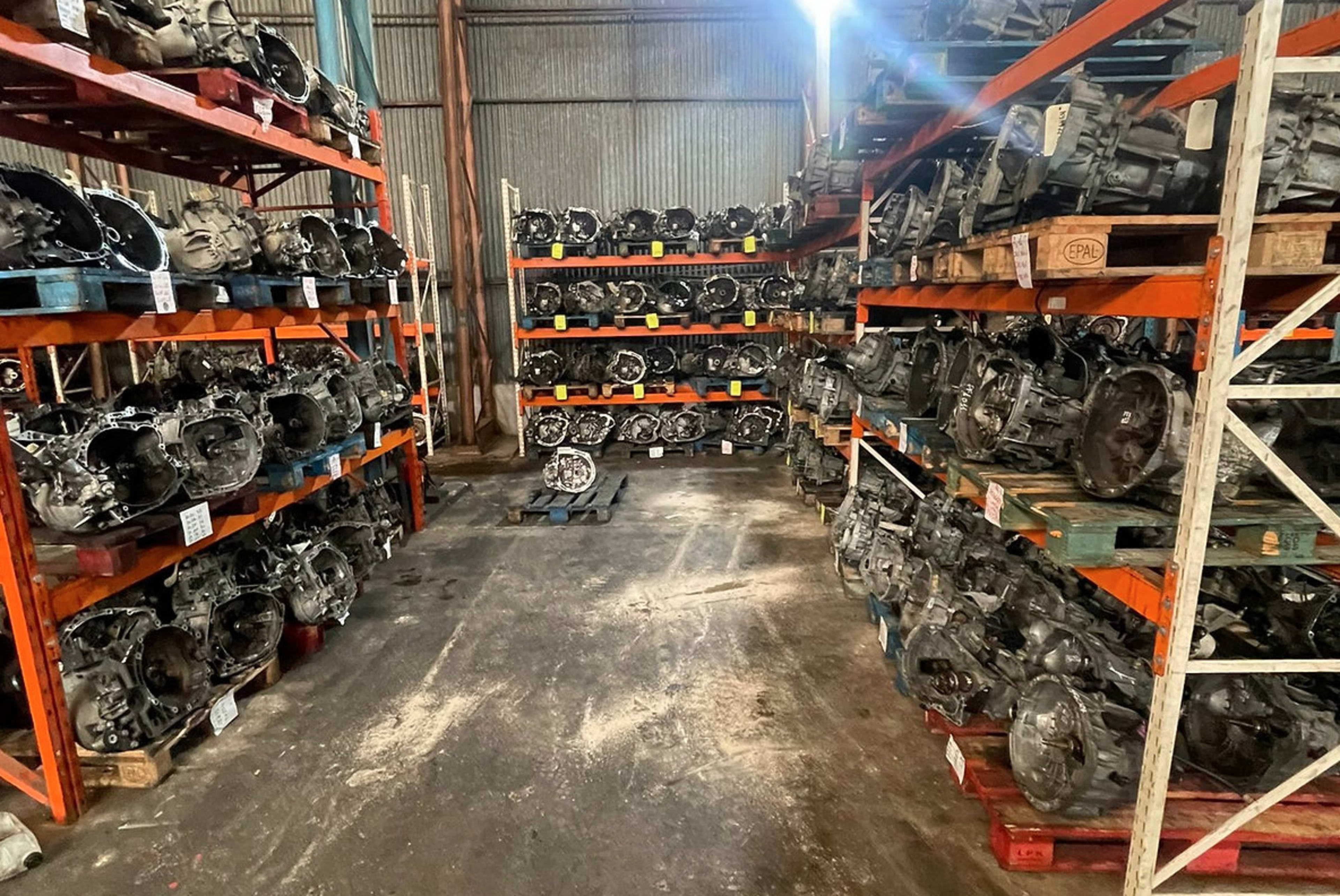 A warehouse aisle lined with metal shelving units filled with various automotive transmissions and engine parts.