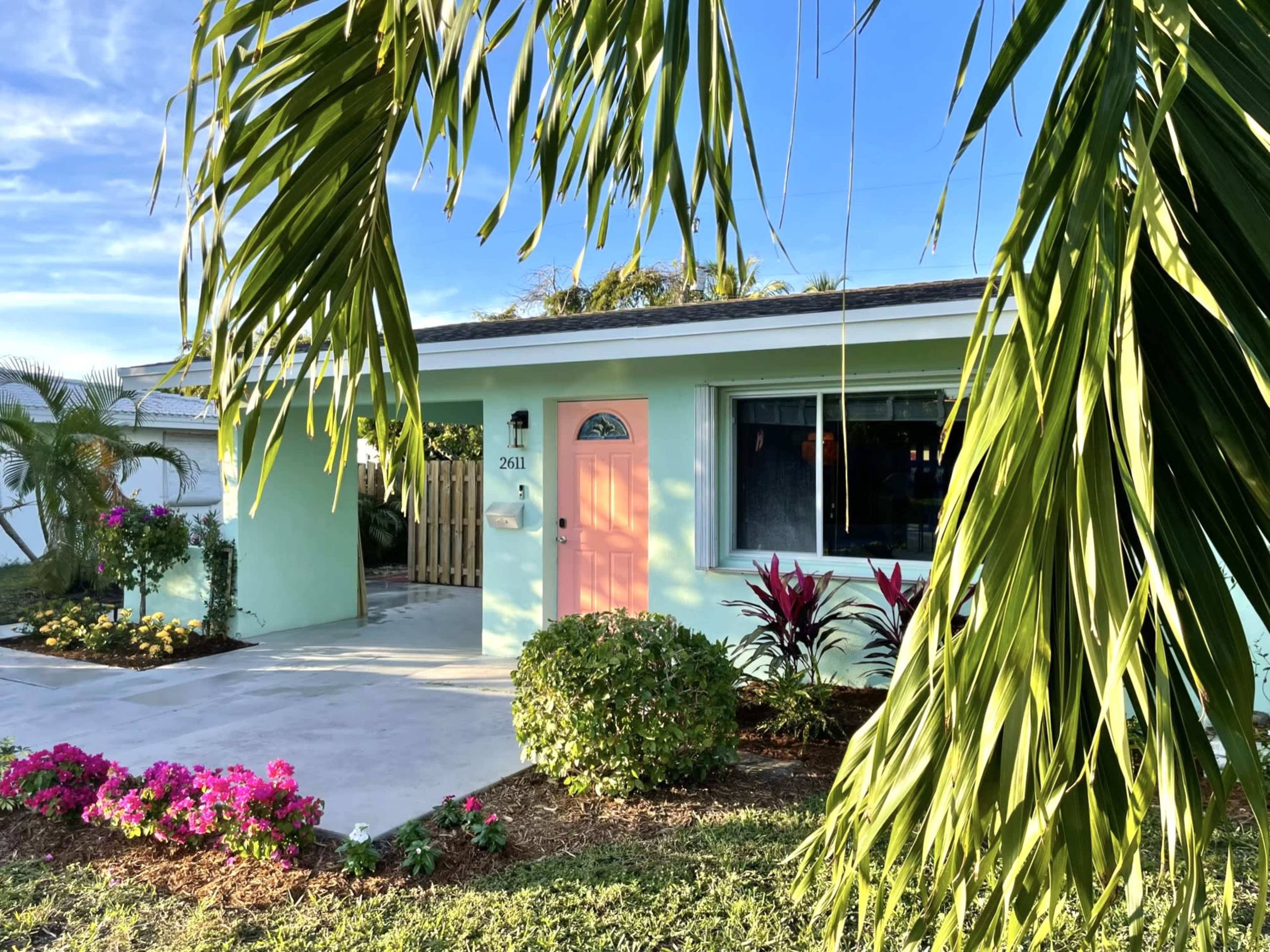 The image shows a light green house with a pink door, surrounded by tropical plants and flowers, under a clear blue sky.