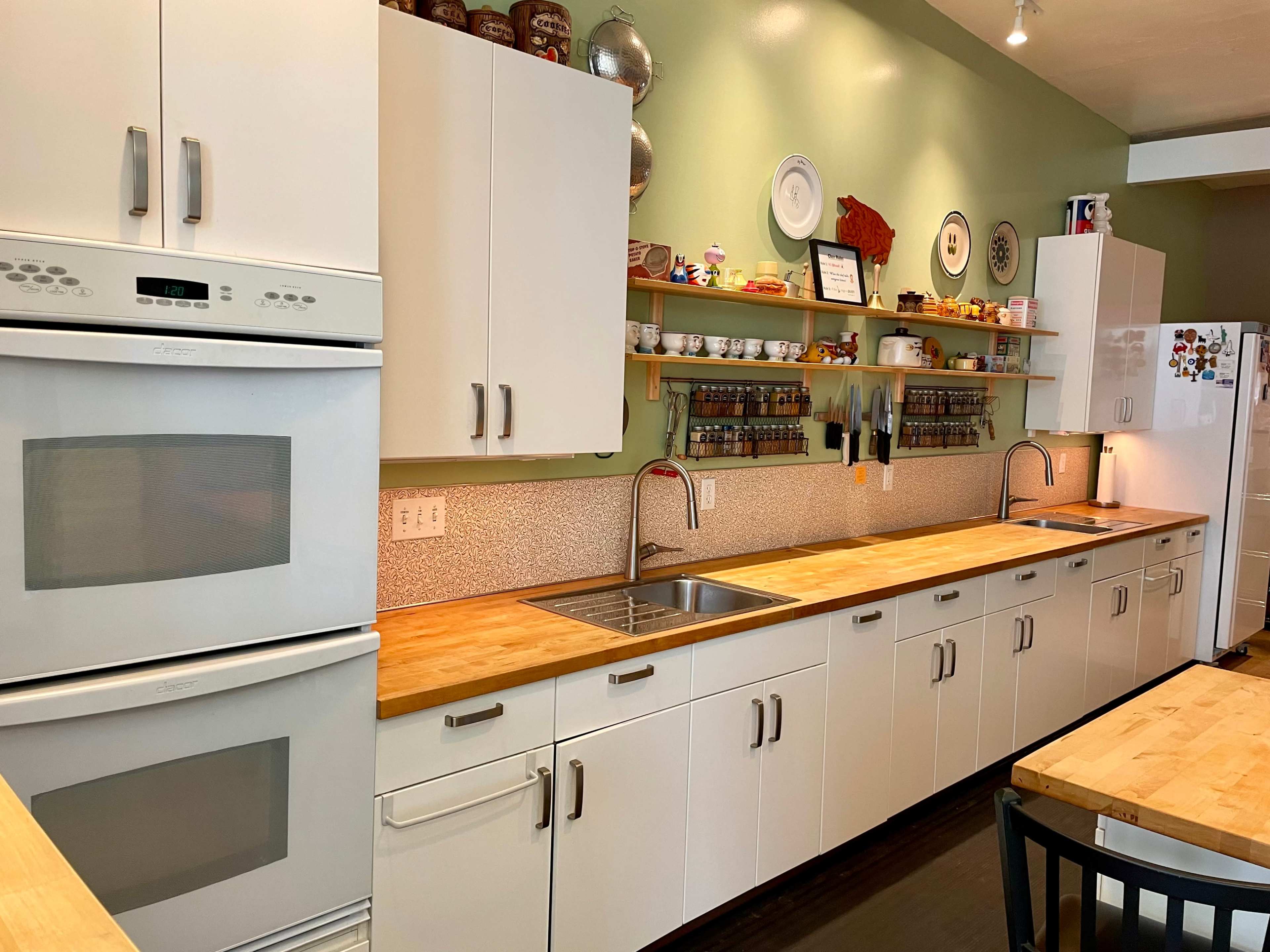 The kitchen features a long wooden countertop with double sinks, white cabinets, and organized shelves displaying dishes and spice jars against a green wall.