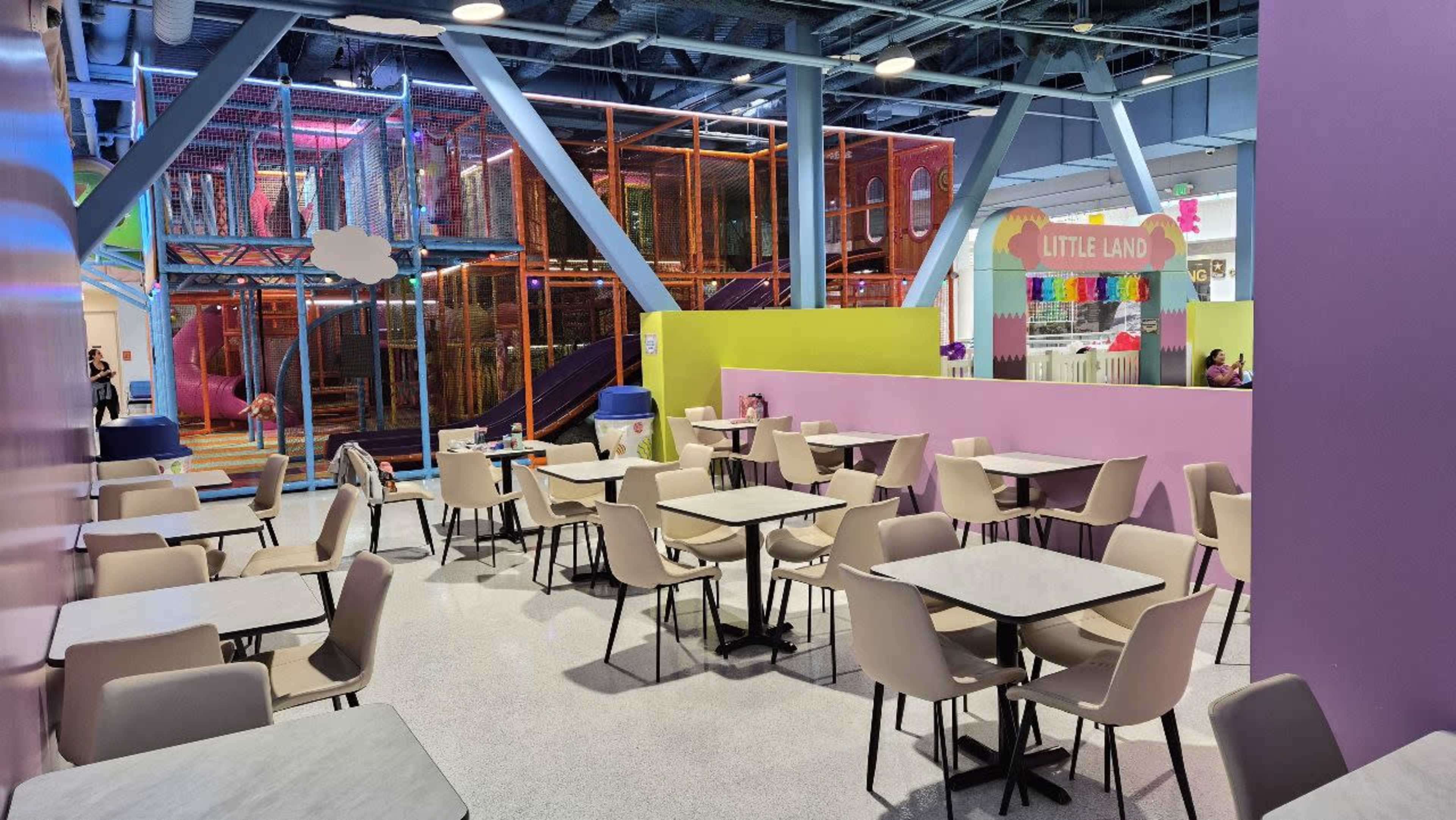 The image shows a colorful indoor play area featuring seating arrangements with light-colored tables and chairs, alongside a play structure in the background labeled "Little Land."
