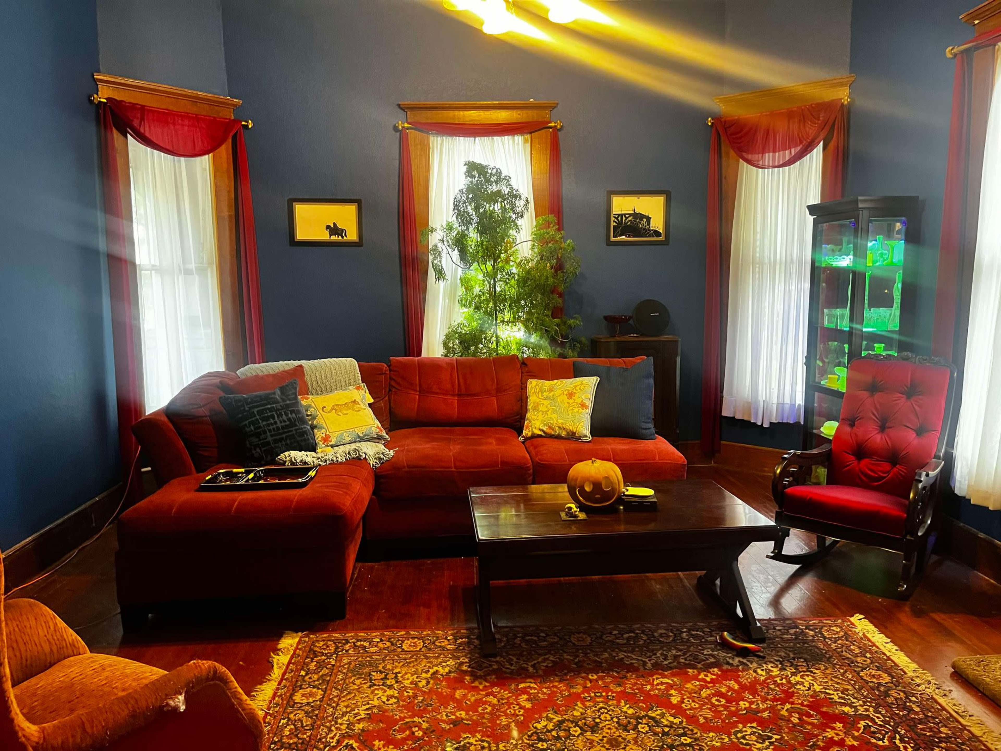 The image shows a cozy living room with a red sectional sofa, a coffee table, and large windows adorned with cream curtains.